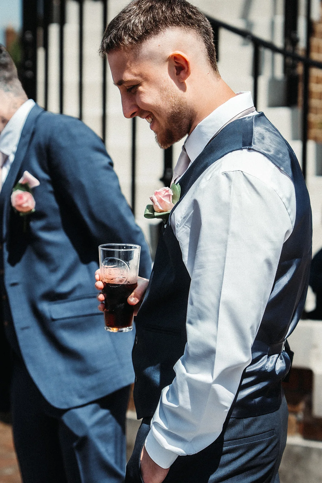A smiling groom wearing a waistcoat and holding a pint glass stands outdoors during the wedding drinks reception alongside a groomsman.