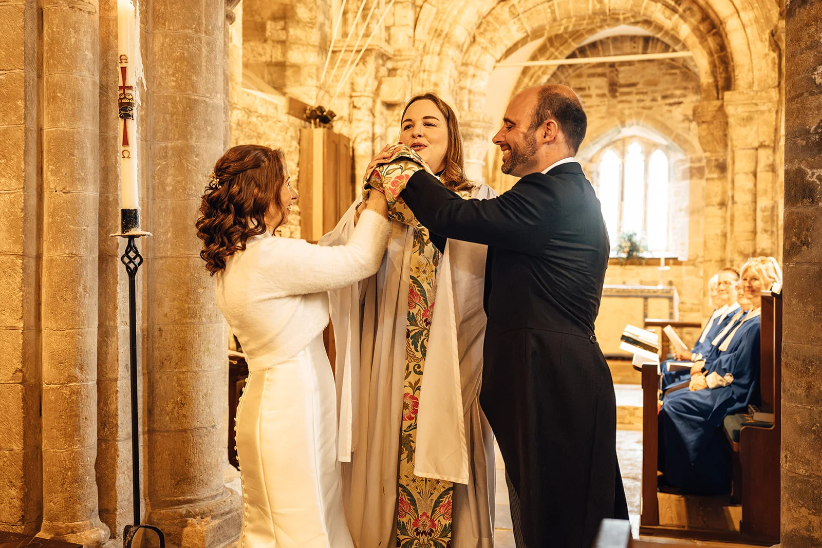 A bride in a satin gown and a groom in a black suit share a joyful moment with the celebrant inside a historic stone church.