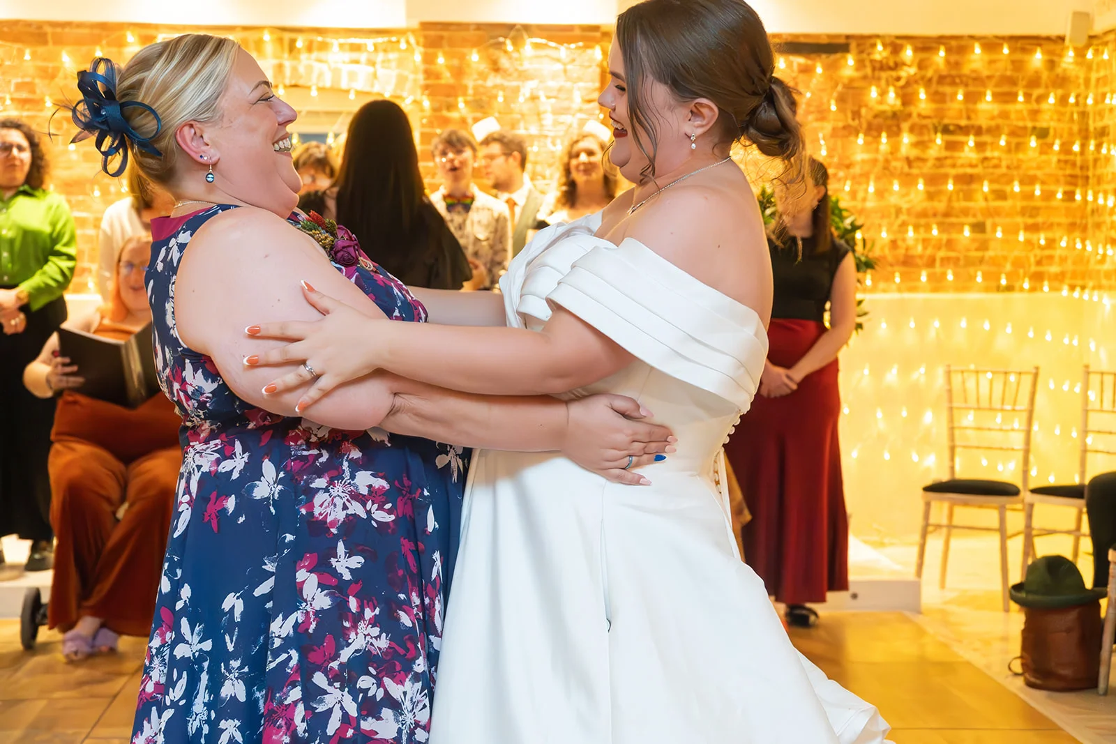 Two smiling brides share a joyful moment dancing together in front of an exposed brick wall illuminated by a warm fairy light canopy.