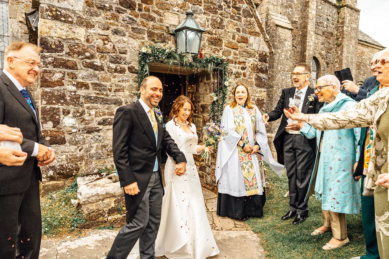 A joyful newlywed couple walks hand in hand out of a stone church doorway as cheering guests shower them with confetti.