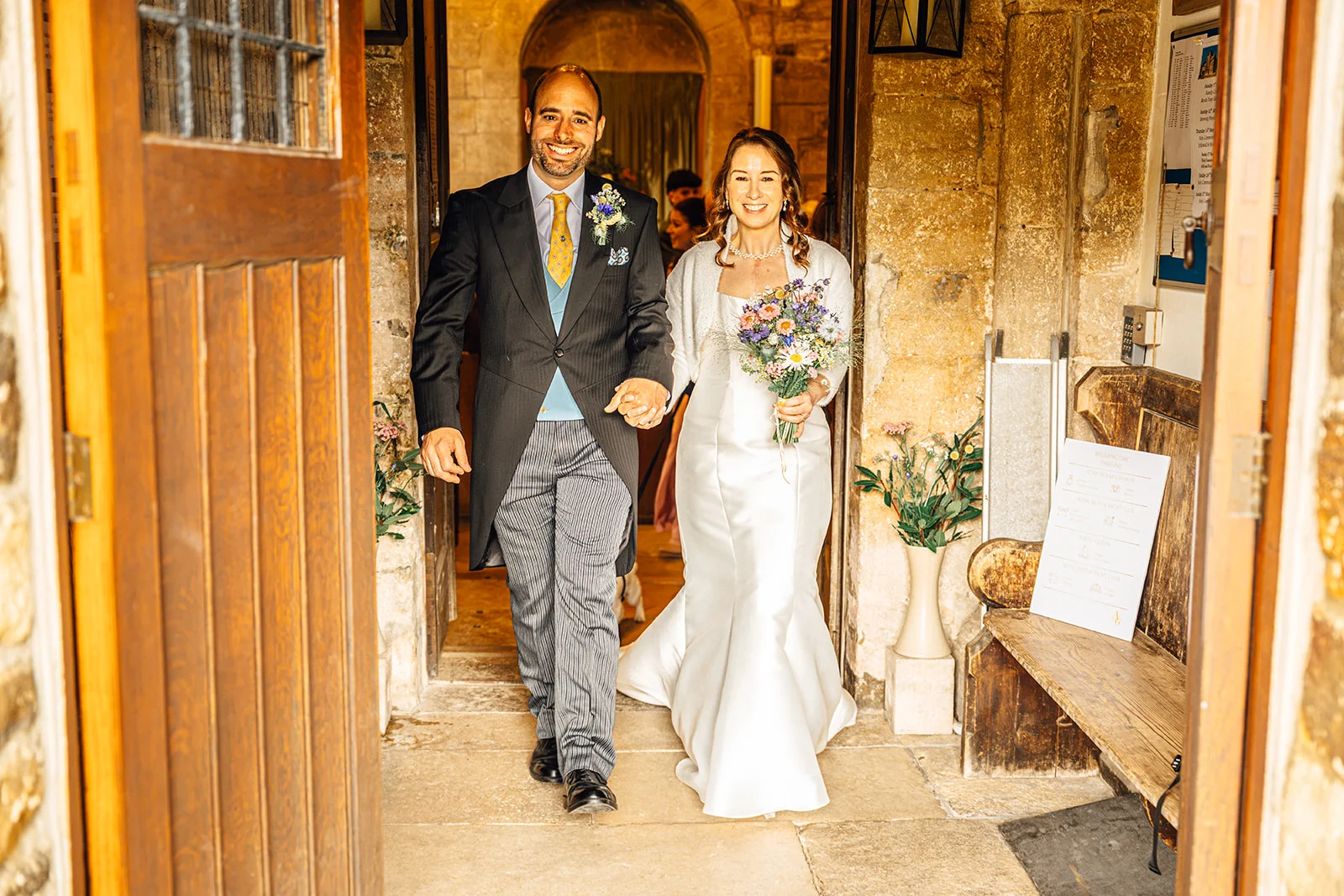 A smiling bride and groom step out of a stone church doorway hand in hand, with the groom wearing a morning suit and the bride in a satin gown holding a wildflower bouquet.