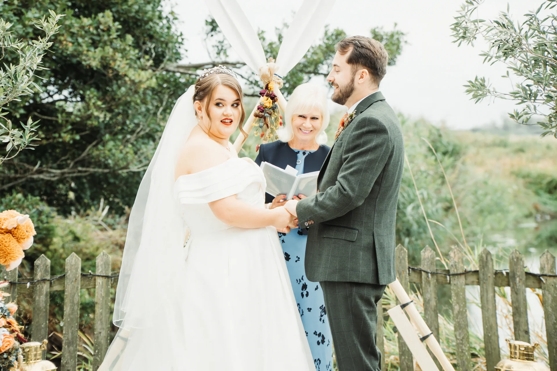A joyful bride and groom hold hands during an outdoor celebrant-led wedding ceremony in front of a wooden fence and greenery.