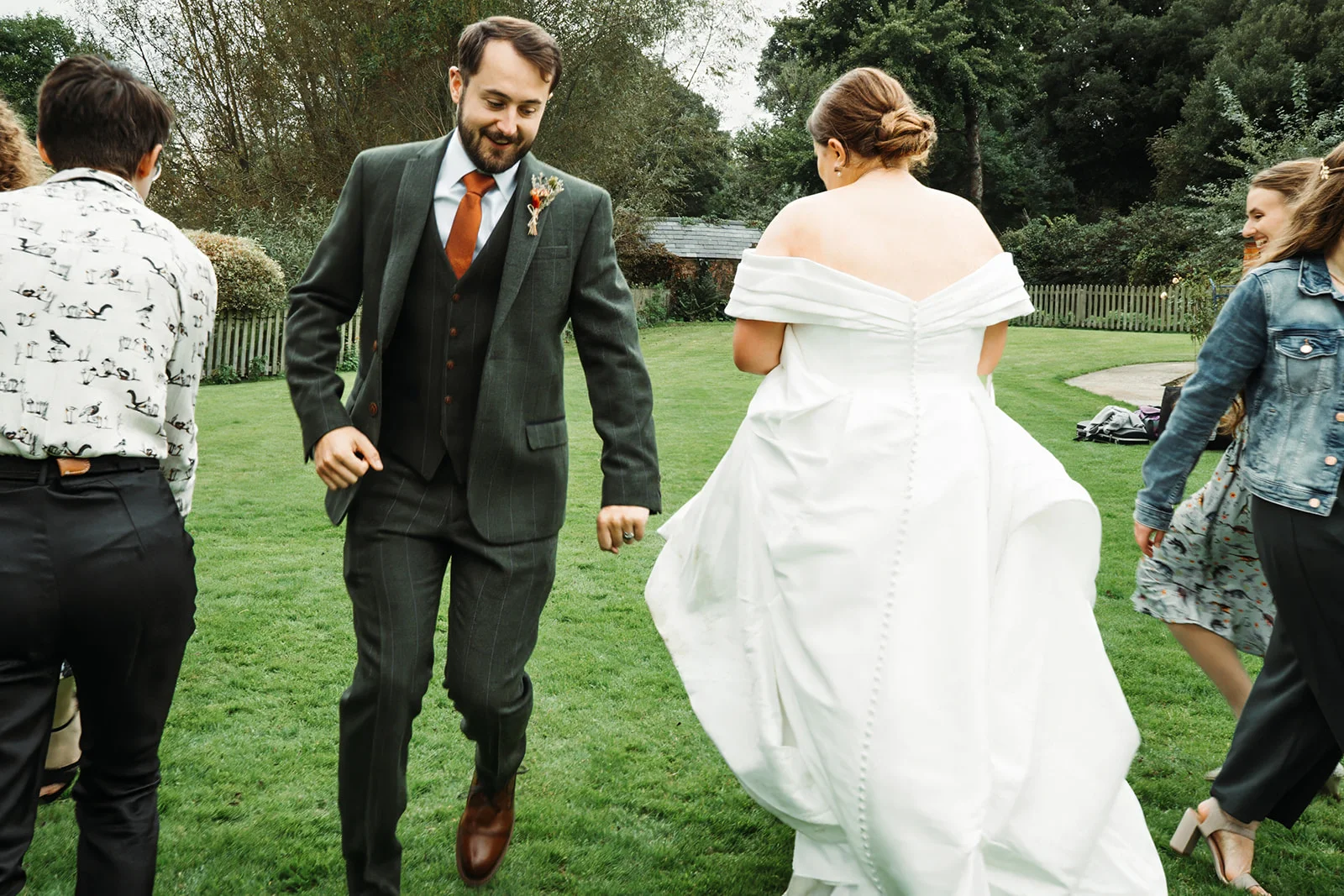 A newlywed couple and their wedding party joyfully run across a lush green lawn during an outdoor wedding celebration.
