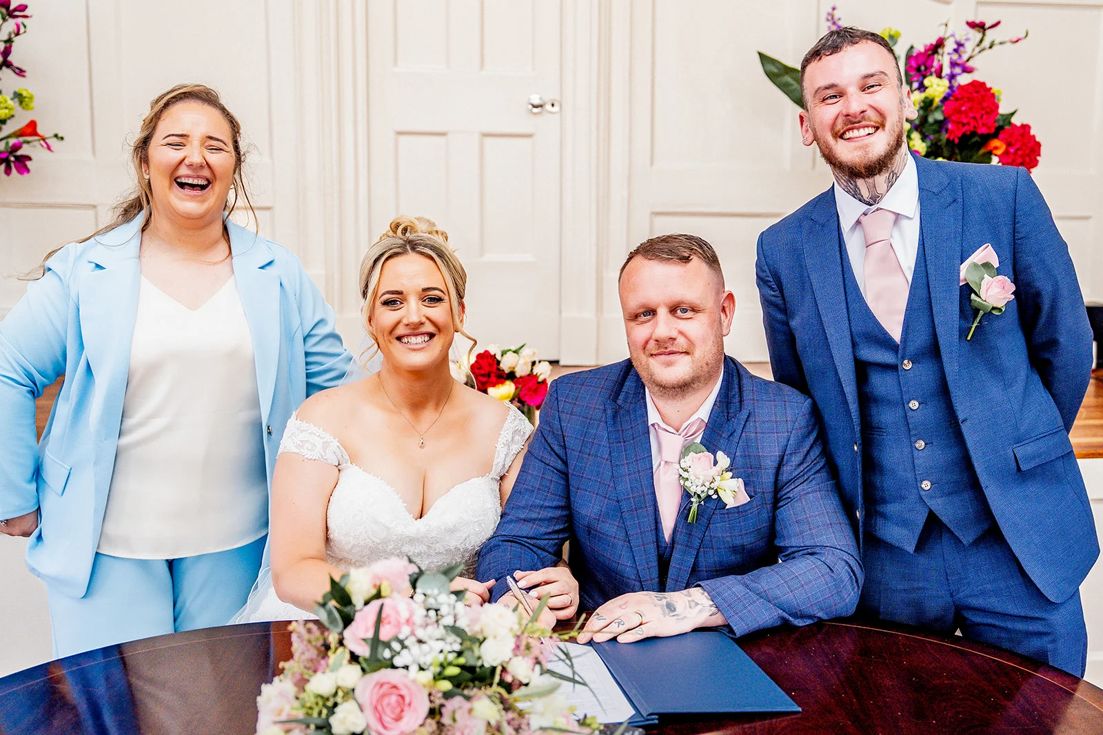 The bride and groom smile while signing their marriage register, accompanied by joyful wedding party members standing behind them in a bright, panelled room.