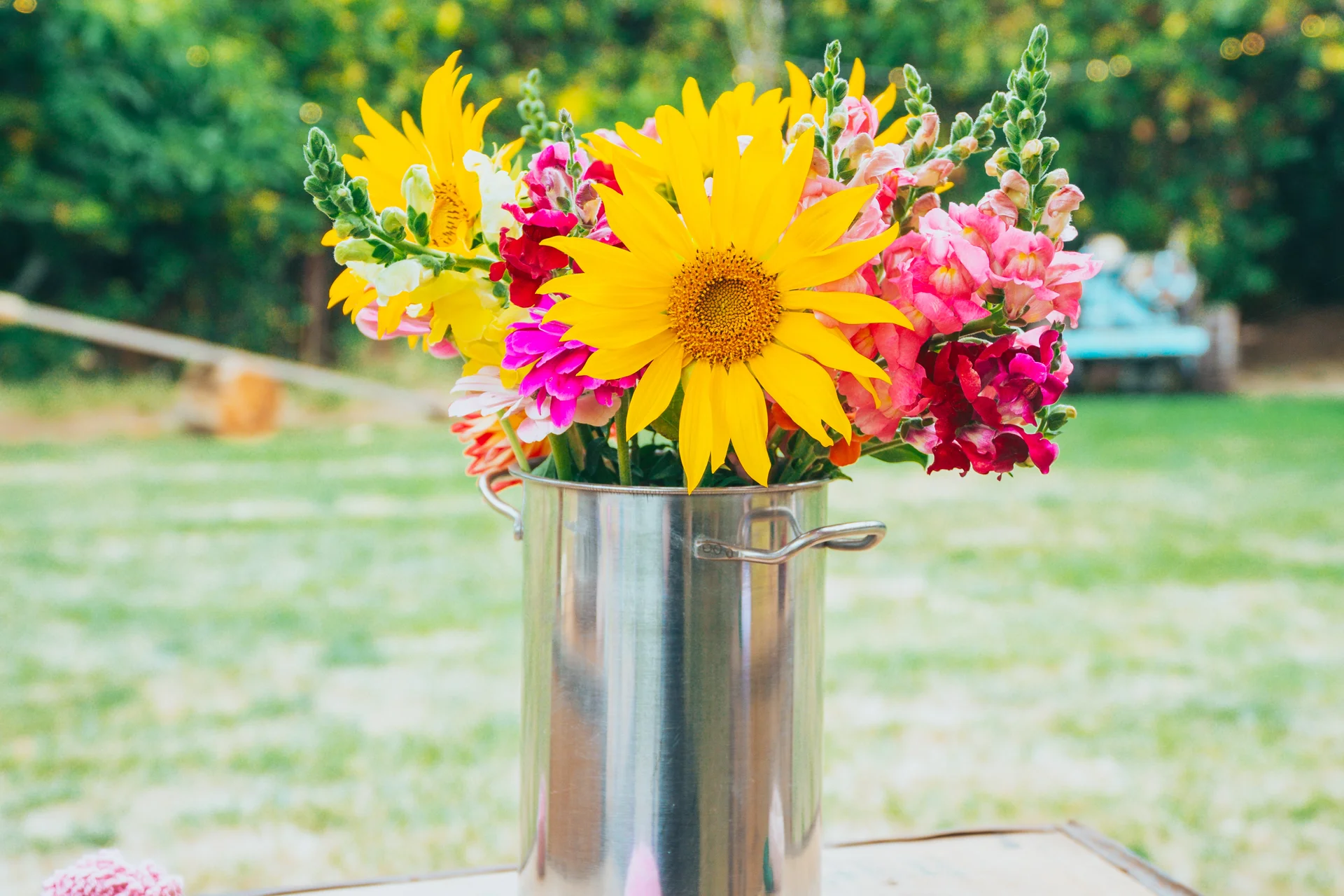 A vibrant wedding centrepiece features a colourful arrangement of bright yellow sunflowers and pink snapdragons displayed in a metal urn outdoors.
