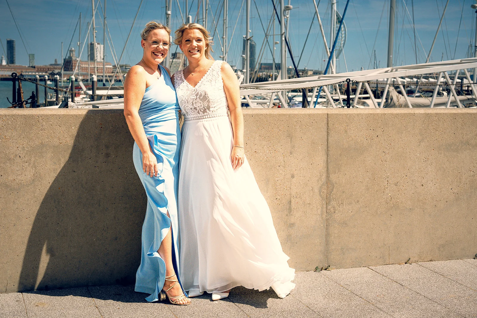 Two smiling brides stand side by side posing in front of a sunny harbour backdrop with boats and masts.
