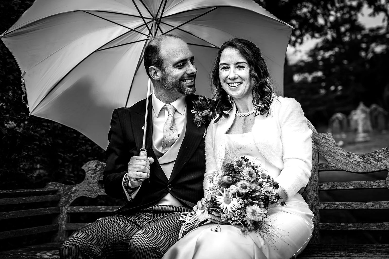 A happily smiling bride and groom sit closely together on an outdoor wooden bench while sheltered beneath a large white umbrella.