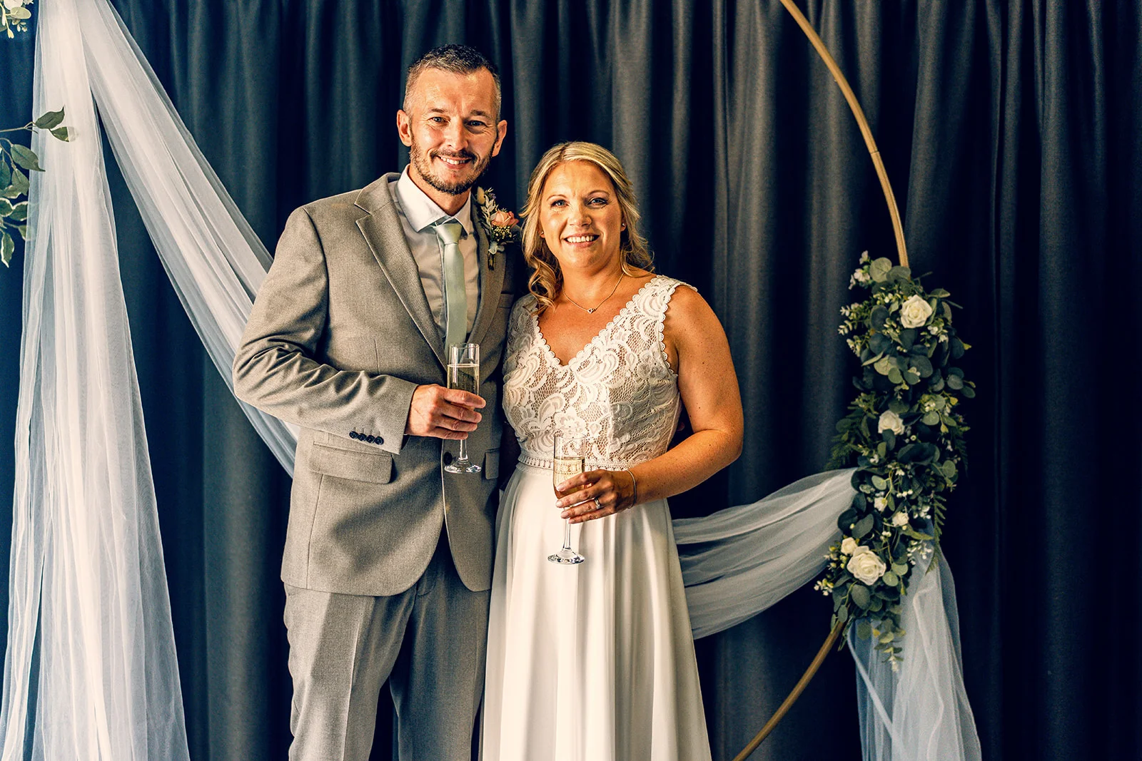 A smiling bride in a lace-trimmed white gown and a groom in a grey suit pose together holding champagne glasses in front of a circular floral arch.