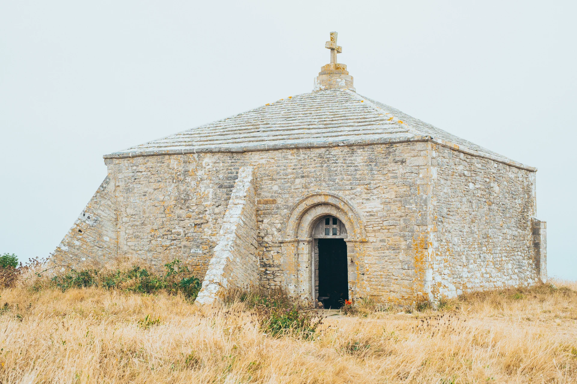 An ancient stone chapel with a pitched roof and a cross on top sits amidst dry grass under an overcast sky.