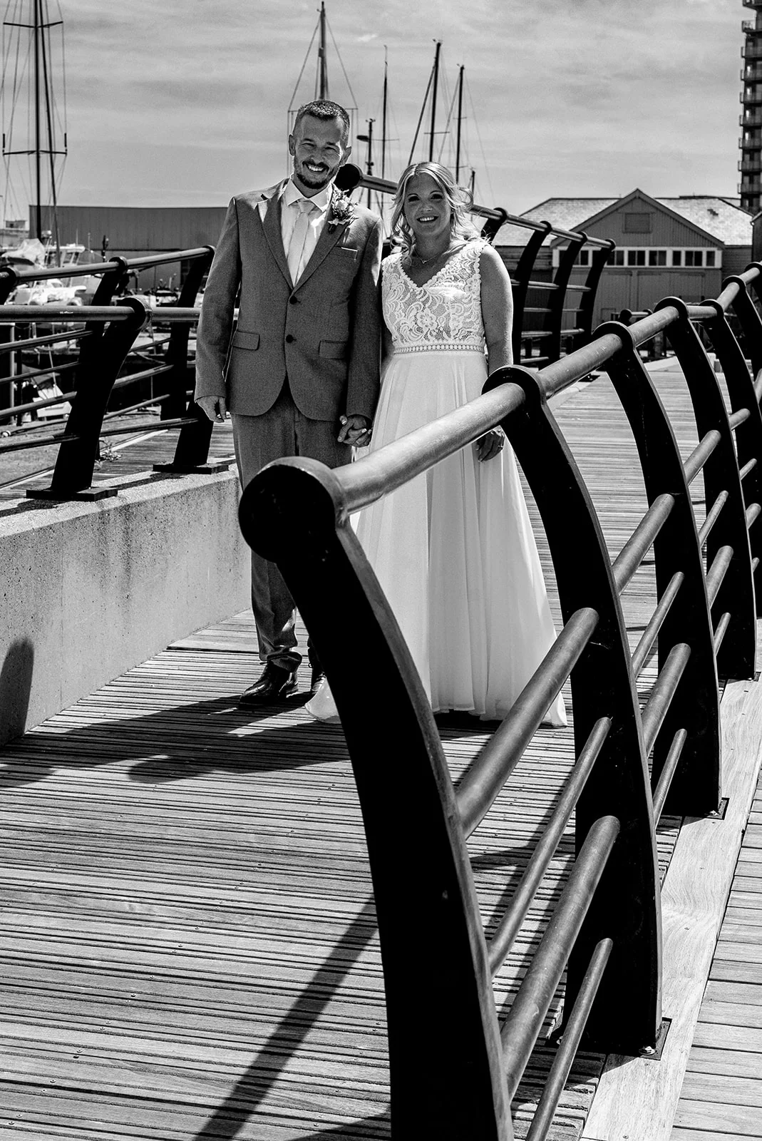 A newlywed couple poses smiling on a wooden pier with a harbour and boat masts visible in the background.