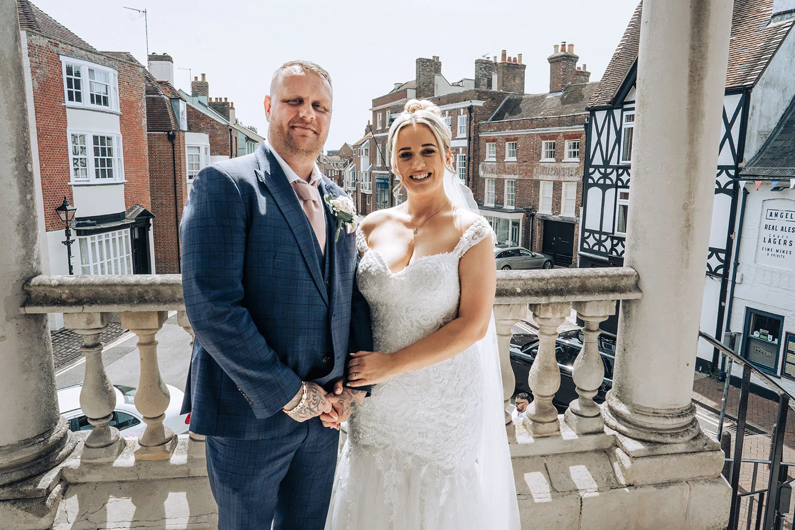 A smiling bride and groom stand together on a stone balcony overlooking a historic town street during their wedding day.
