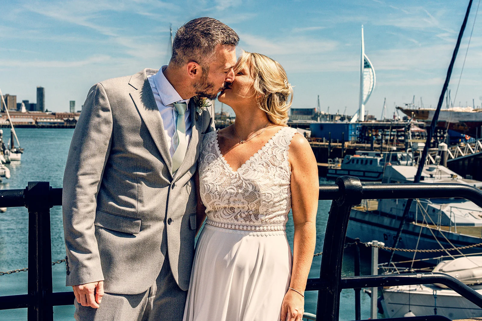 A newlywed couple shares a kiss against a sunny harbour backdrop featuring moored boats and a distinctive modern tower.