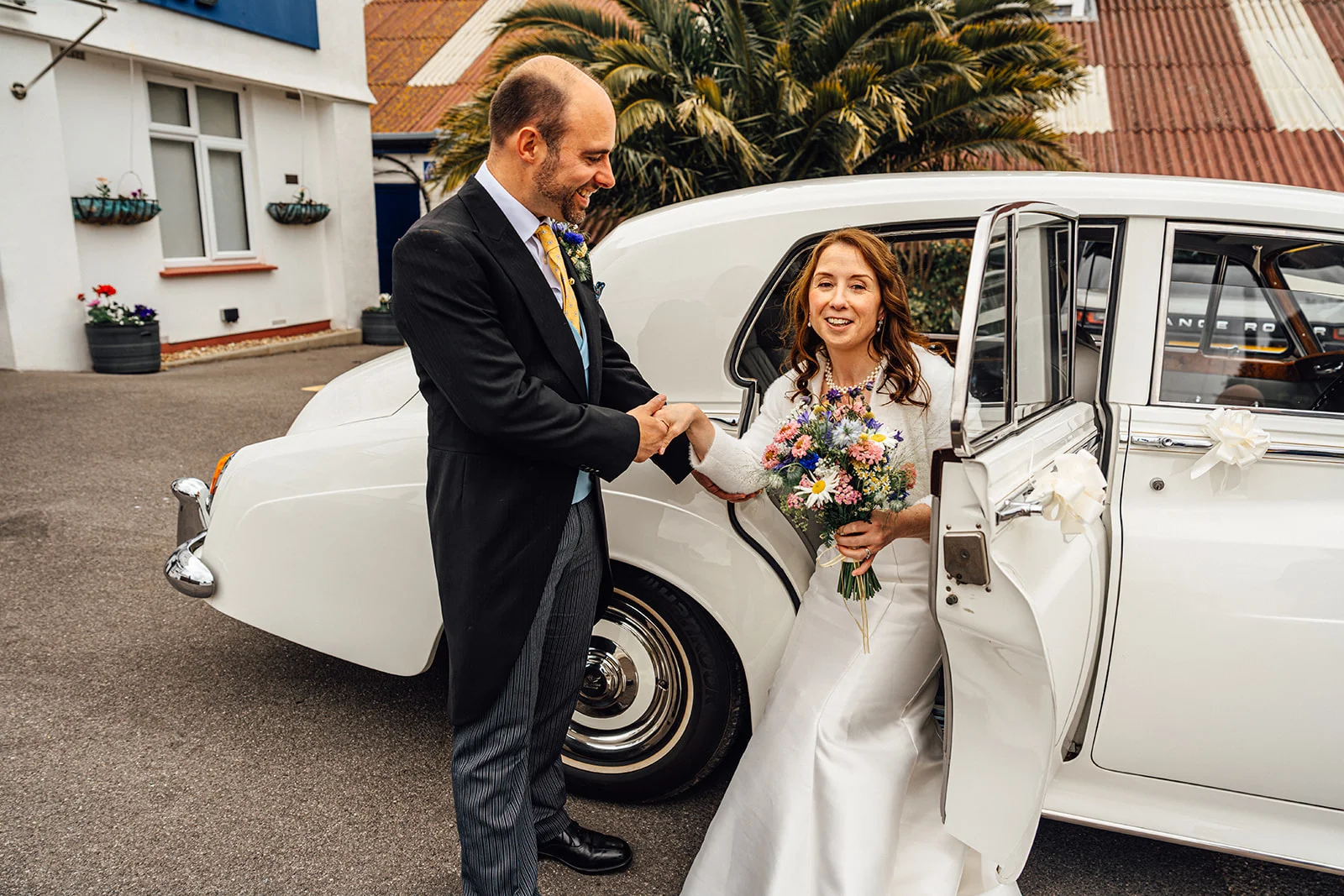 A smiling groom helps the bride out of a classic white wedding car as she holds a colourful wildflower bouquet.