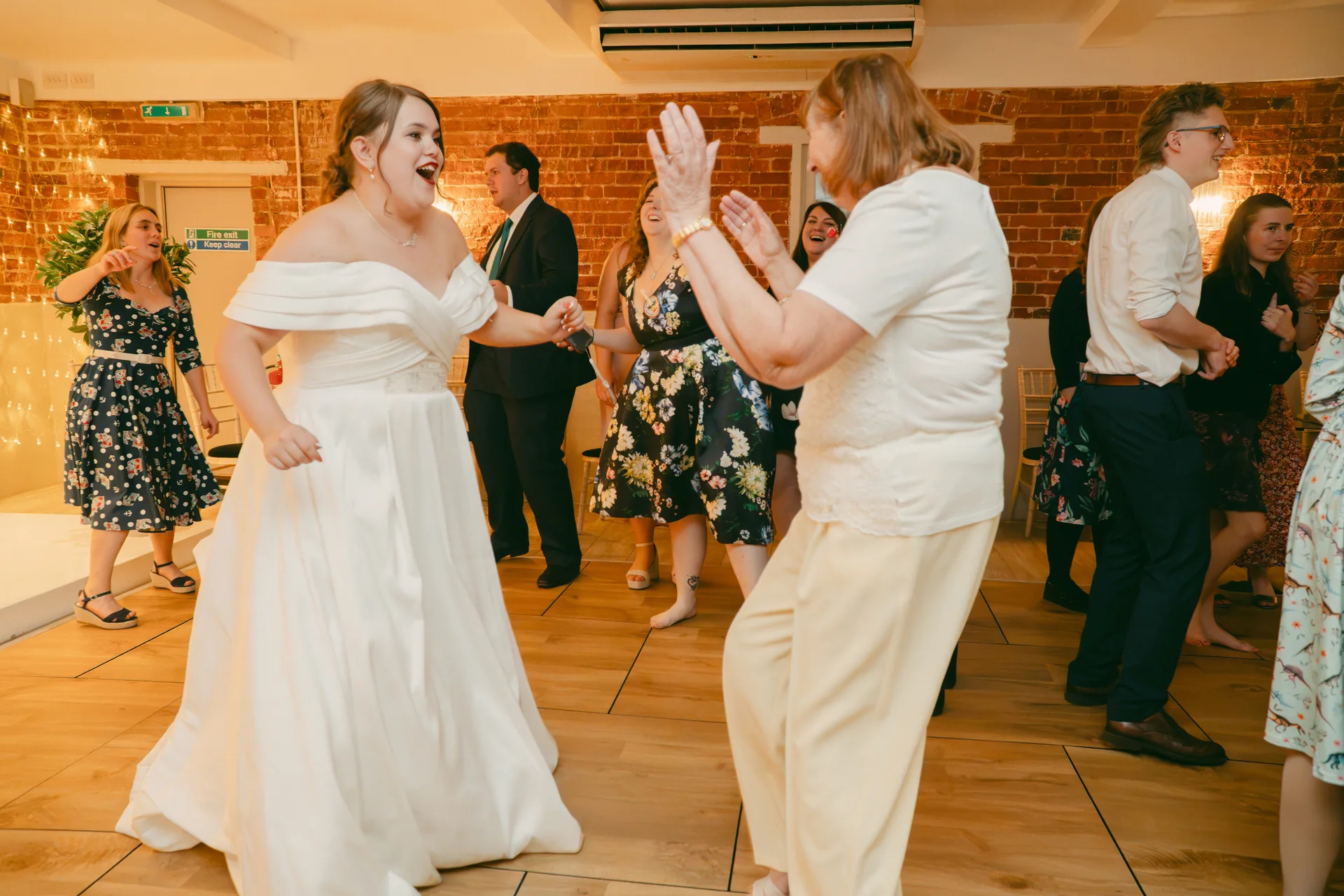 A joyful bride in an off-the-shoulder satin gown laughs and dances enthusiastically on the dance floor surrounded by cheering guests in a rustic venue with exposed brick walls.