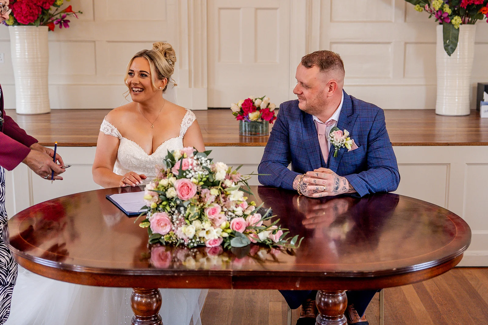A smiling bride in an off-the-shoulder lace dress and her groom in a blue checked suit sit at a wooden table while signing the register during their civil ceremony.