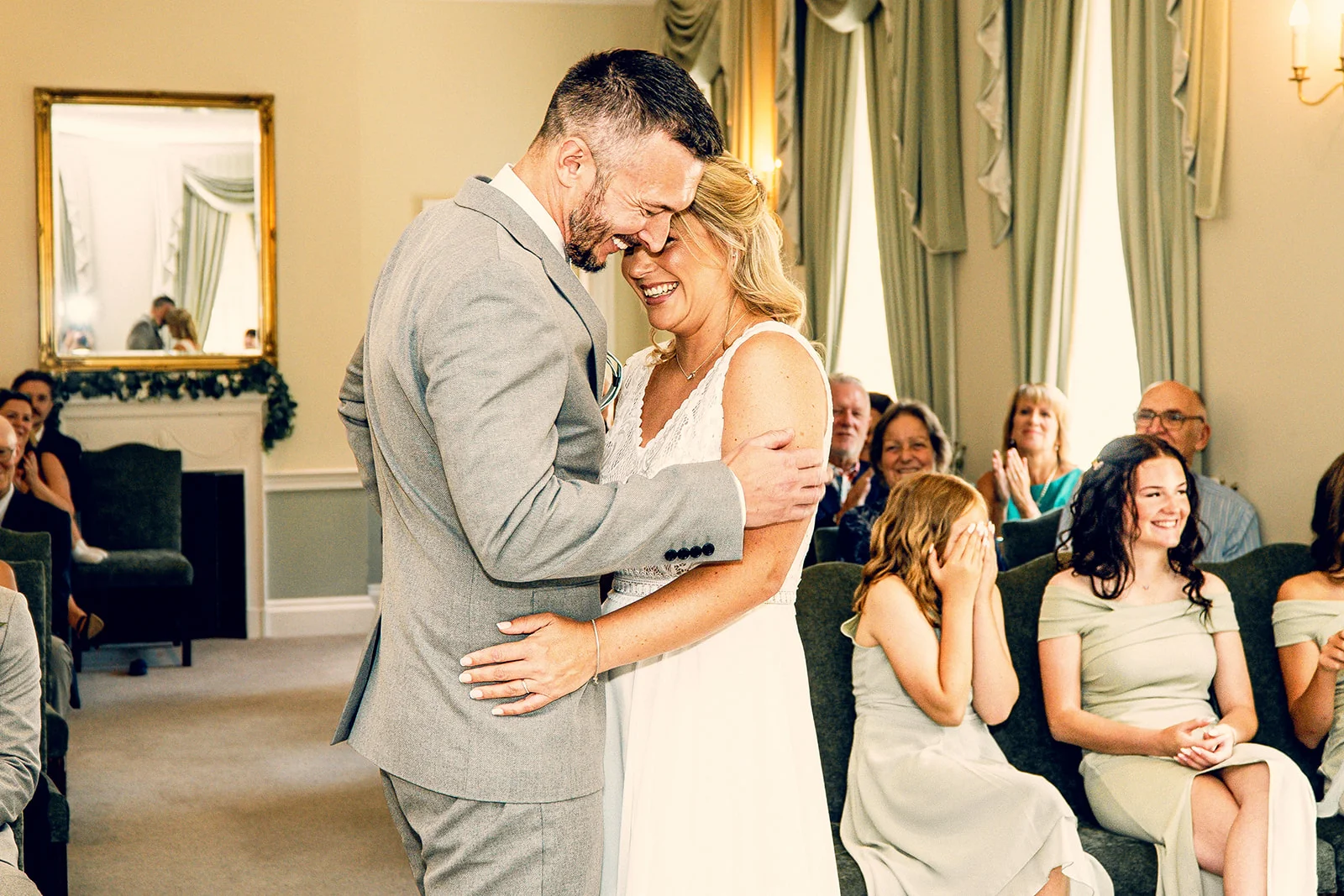 A smiling bride and groom embrace joyfully during their indoor ceremony as seated guests look on and laugh.
