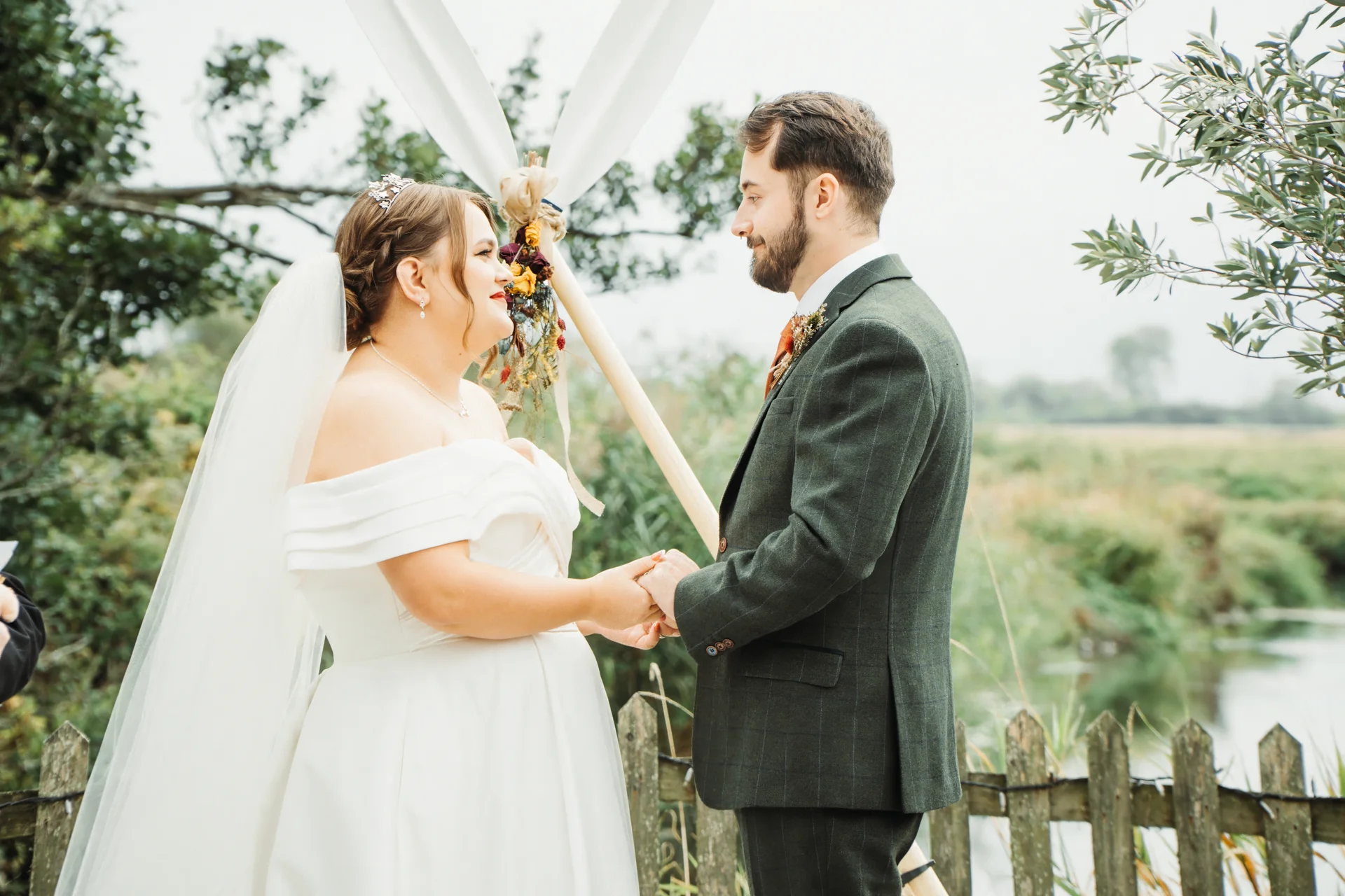A bride in an off-the-shoulder white gown and veil holds hands lovingly with her groom in a green suit during an outdoor wedding ceremony.