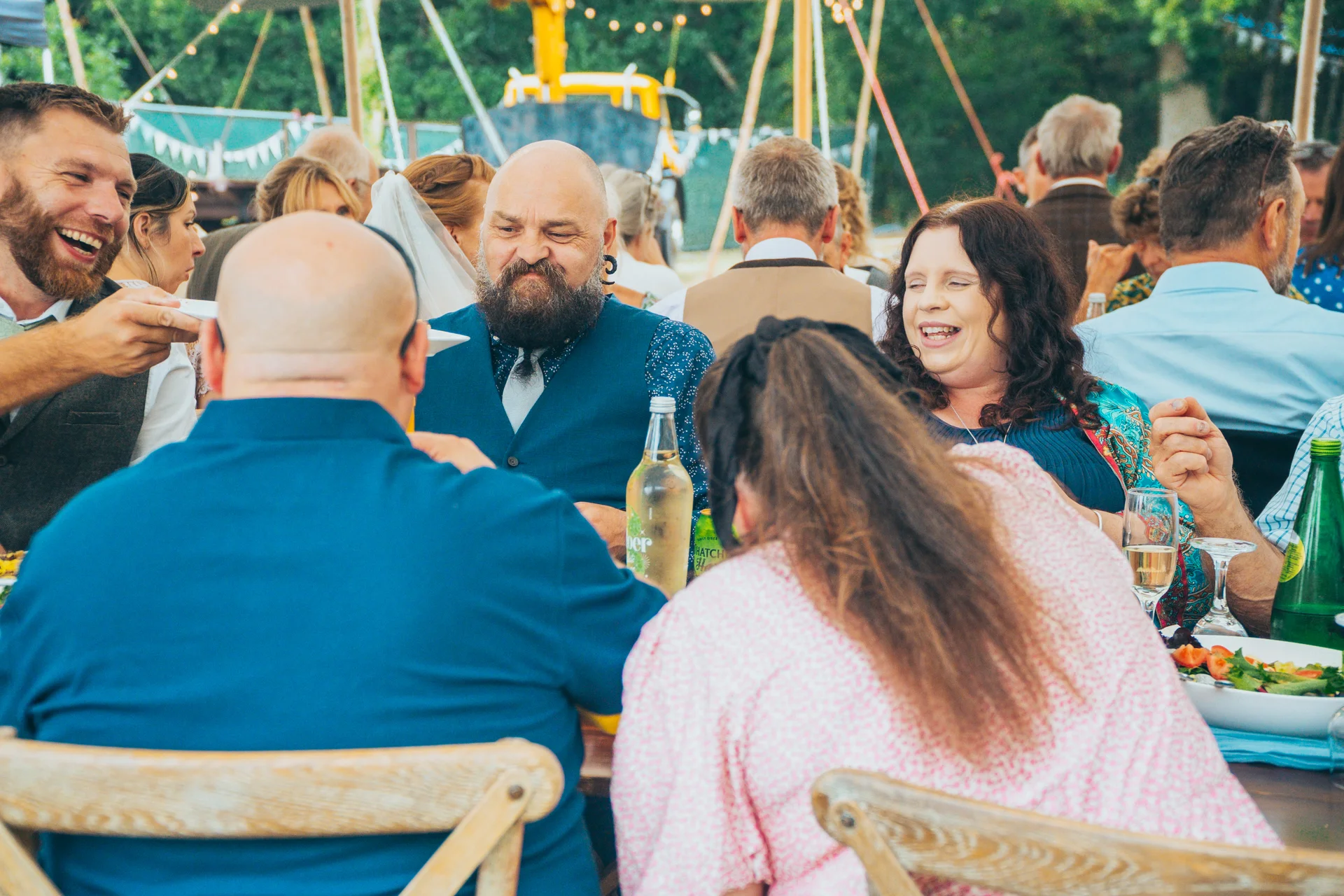 Wedding guests share a joyful, laughter-filled moment around a rustic banquet table inside a brightly lit marquee during the wedding breakfast.