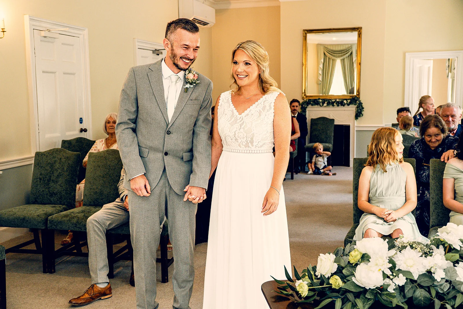 A smiling bride and groom hold hands at the front of a ceremony room while seated guests watch on.