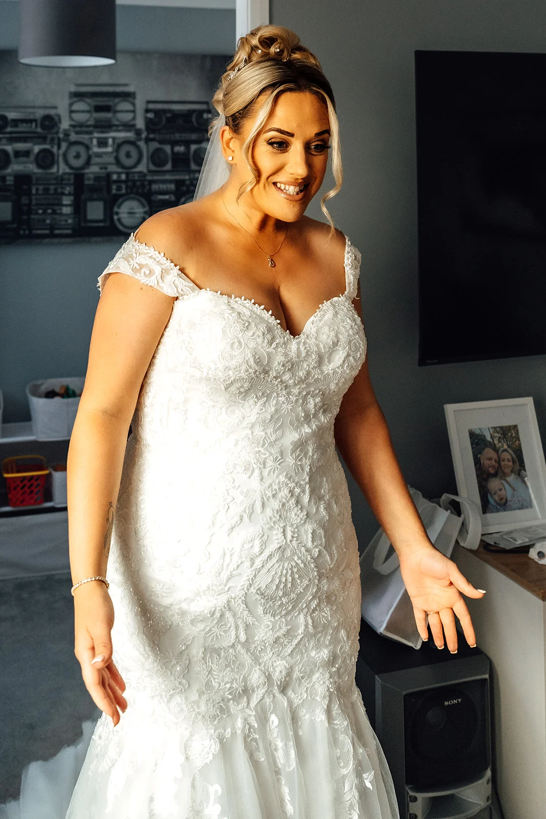 A smiling bride stands indoors during morning preparations, wearing an intricately embroidered lace off-the-shoulder wedding gown and her hair styled in an elegant high bun.