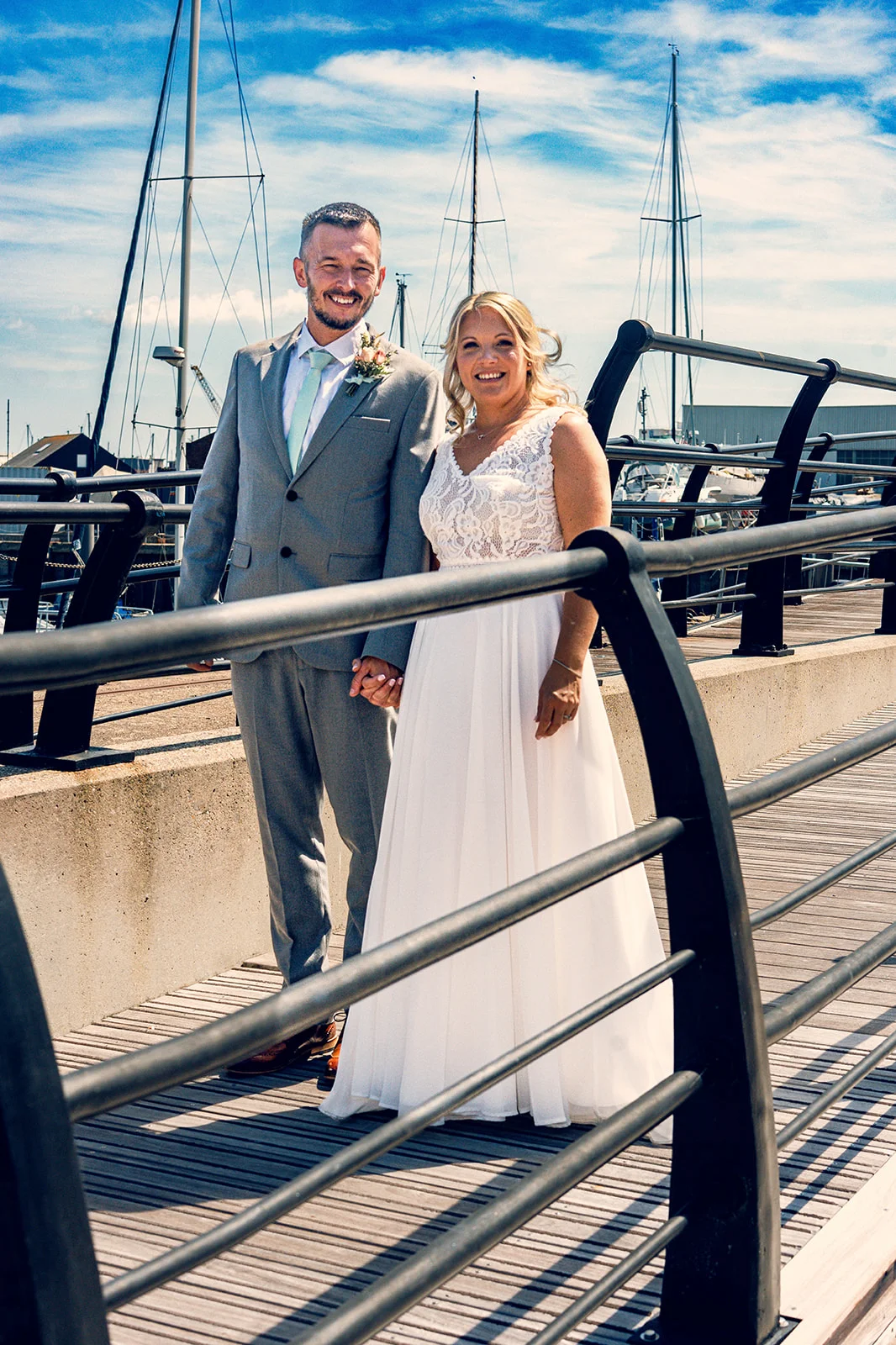 A smiling bride and groom stand holding hands on a wooden pier with a marina and clear blue sky in the background.