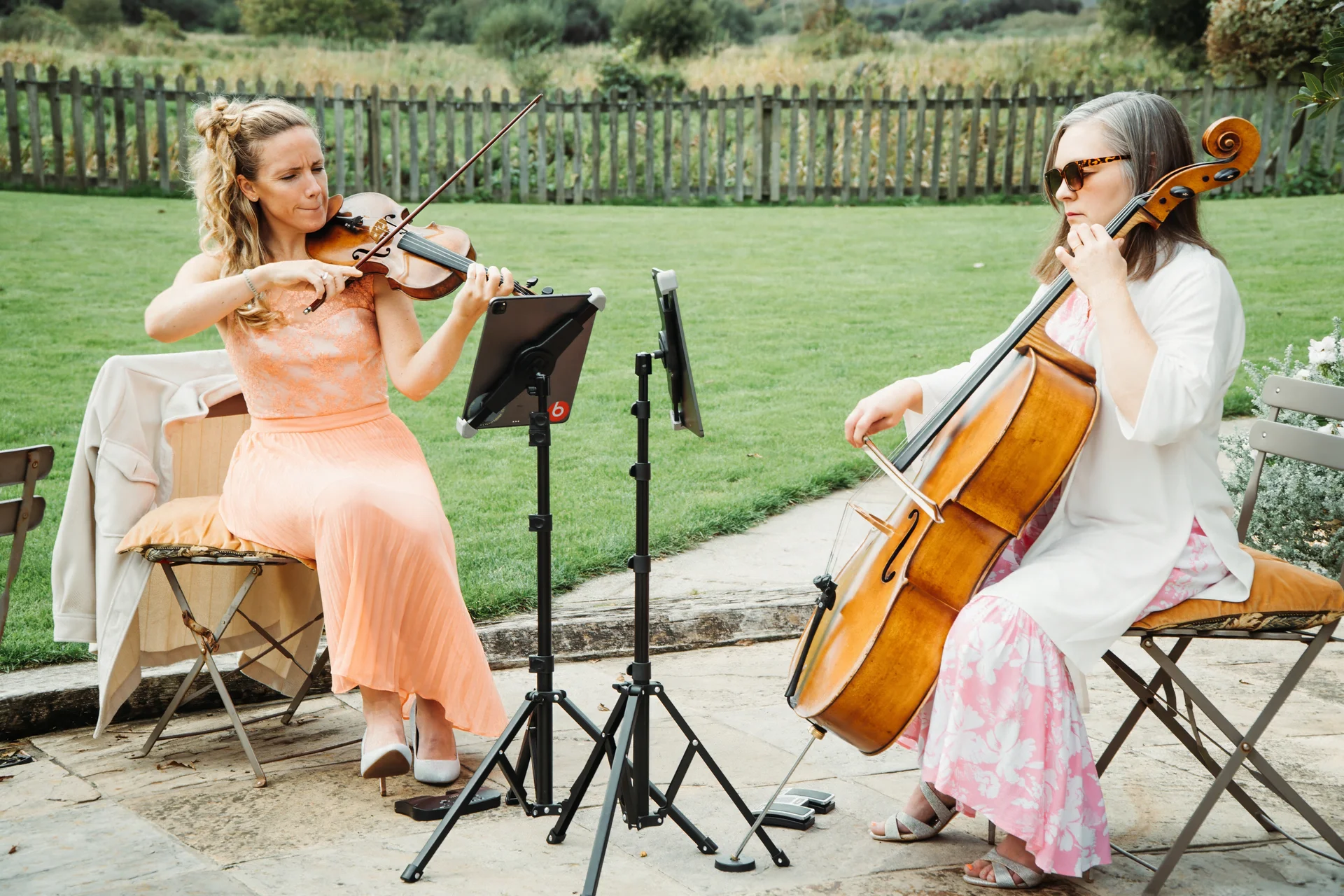 A female violinist in a peach dress and a cellist in a white top play music outdoors on a stone patio during a wedding.