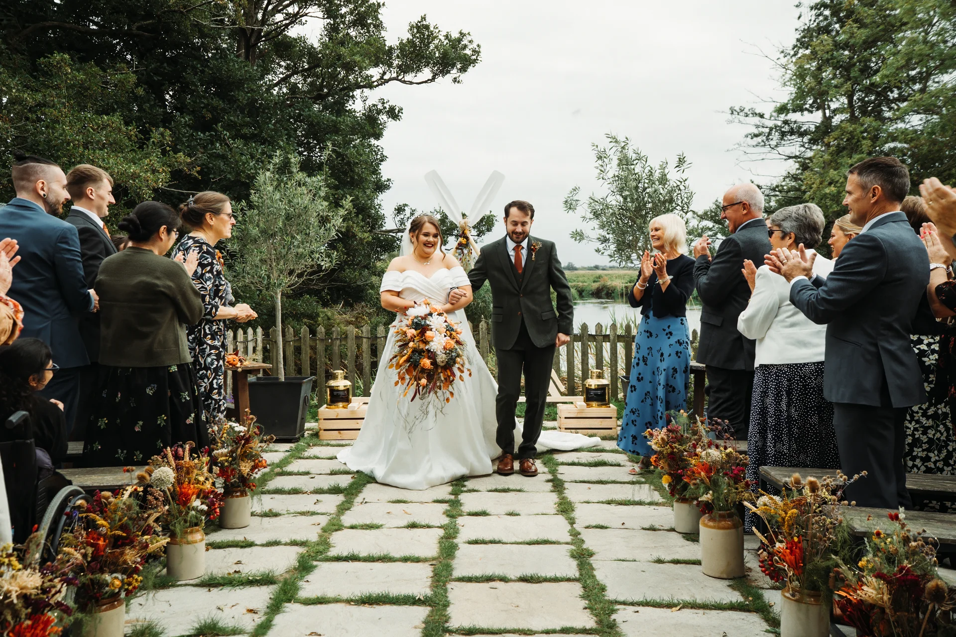 A newlywed couple walks down an outdoor stone aisle hand in hand, smiling as cheering guests applaud them amidst autumnal floral arrangements and a lakeside backdrop.