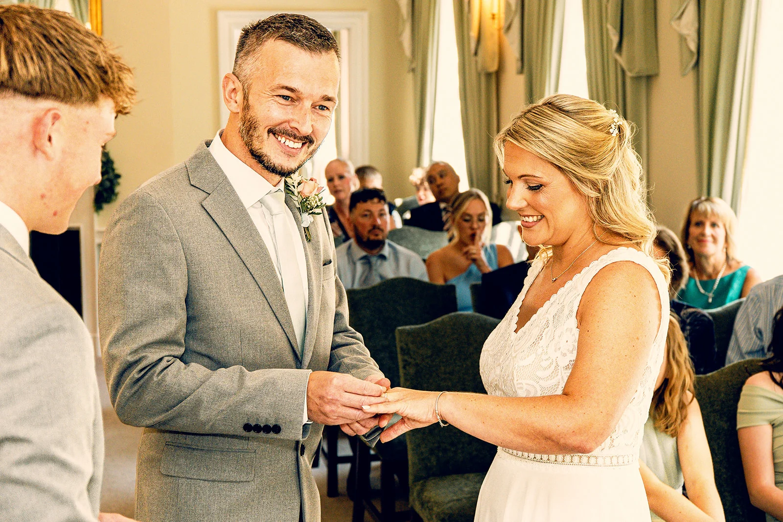 The smiling groom gently places a wedding ring onto the bride's finger during their indoor civil ceremony watched by seated guests.