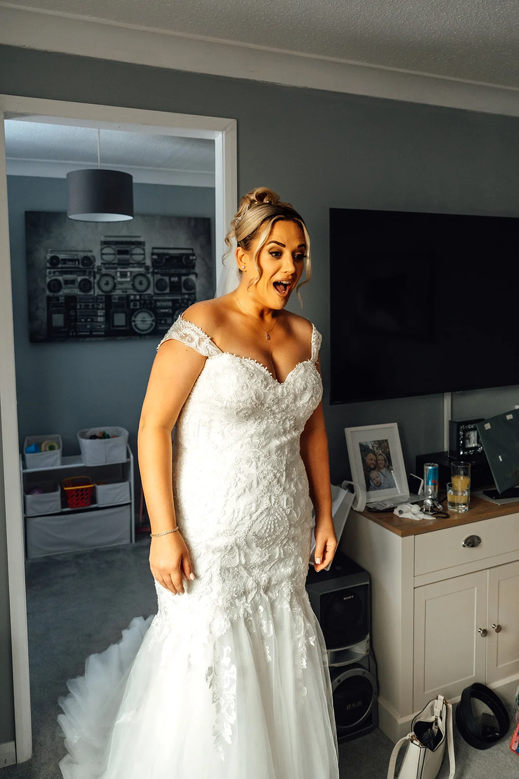 A bride in a lace off-the-shoulder fit and flare gown reacts with an excited expression and open mouth during wedding morning preparations.