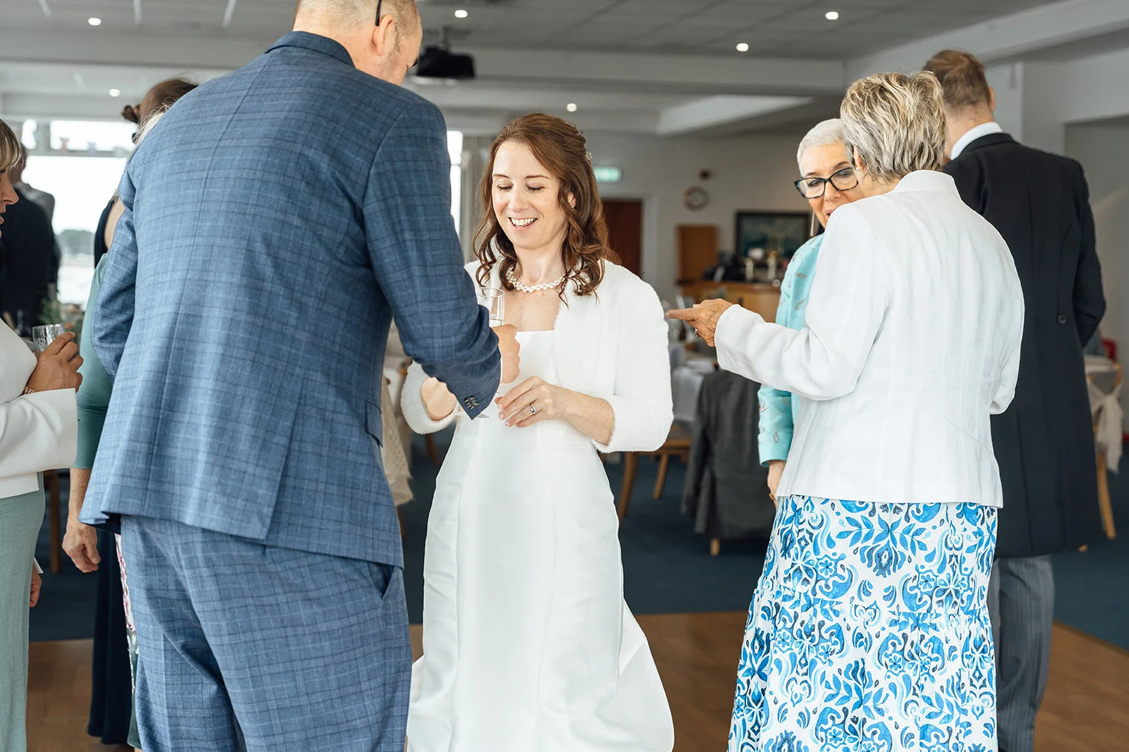 A smiling bride holding a glass during the drinks reception while guests mingle around her in a bright hotel venue.