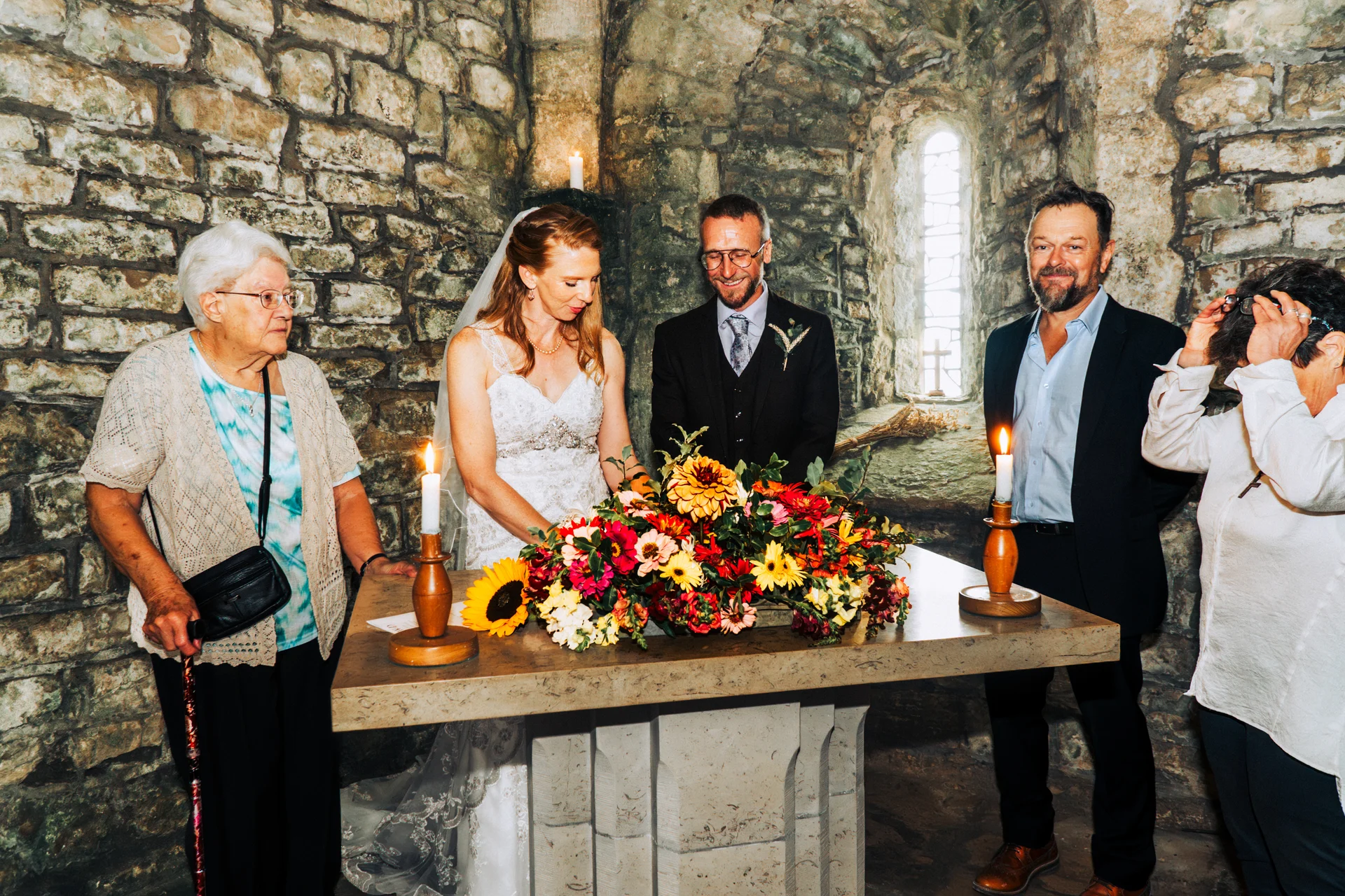 A bride and groom stand at a stone altar adorned with vibrant autumn flowers and lit candles inside a historic stone church, flanked by family members.