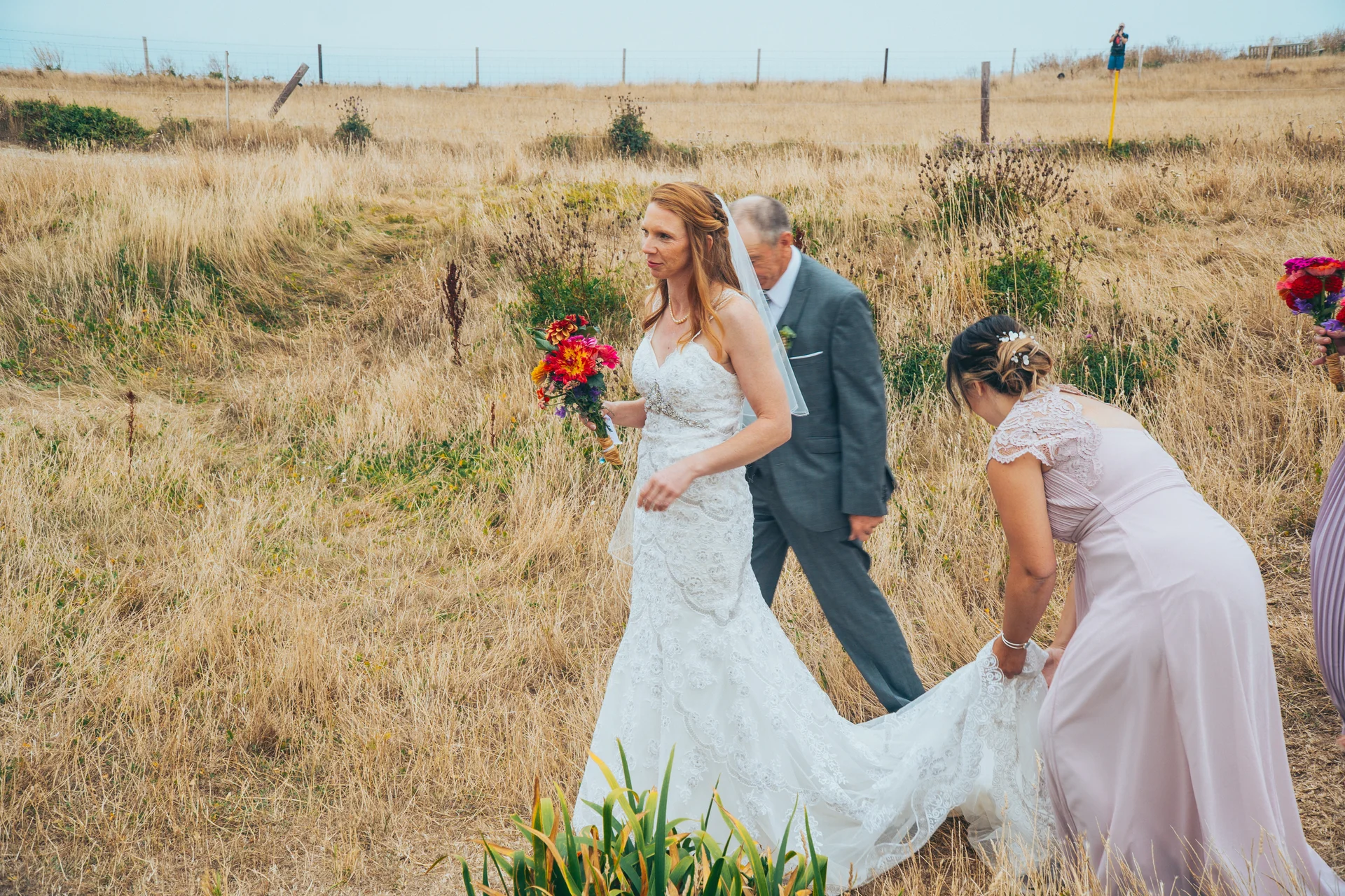 A bride in a lace wedding dress holding a vibrant bouquet walks through a dry grassy field alongside her father, while a bridesmaid carefully holds her train.