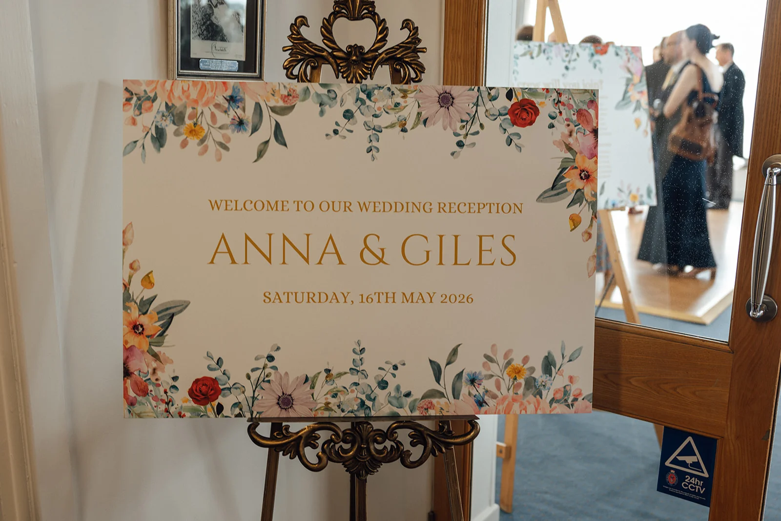 A decorative wedding welcome sign featuring watercolour floral illustrations and gold lettering sits on an ornate stand near a glass doorway.