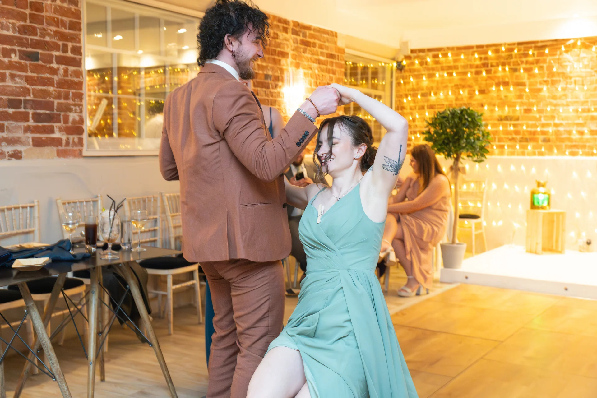 A newlywed couple shares a joyful dance during their evening reception, surrounded by exposed brick walls and warm string lighting.