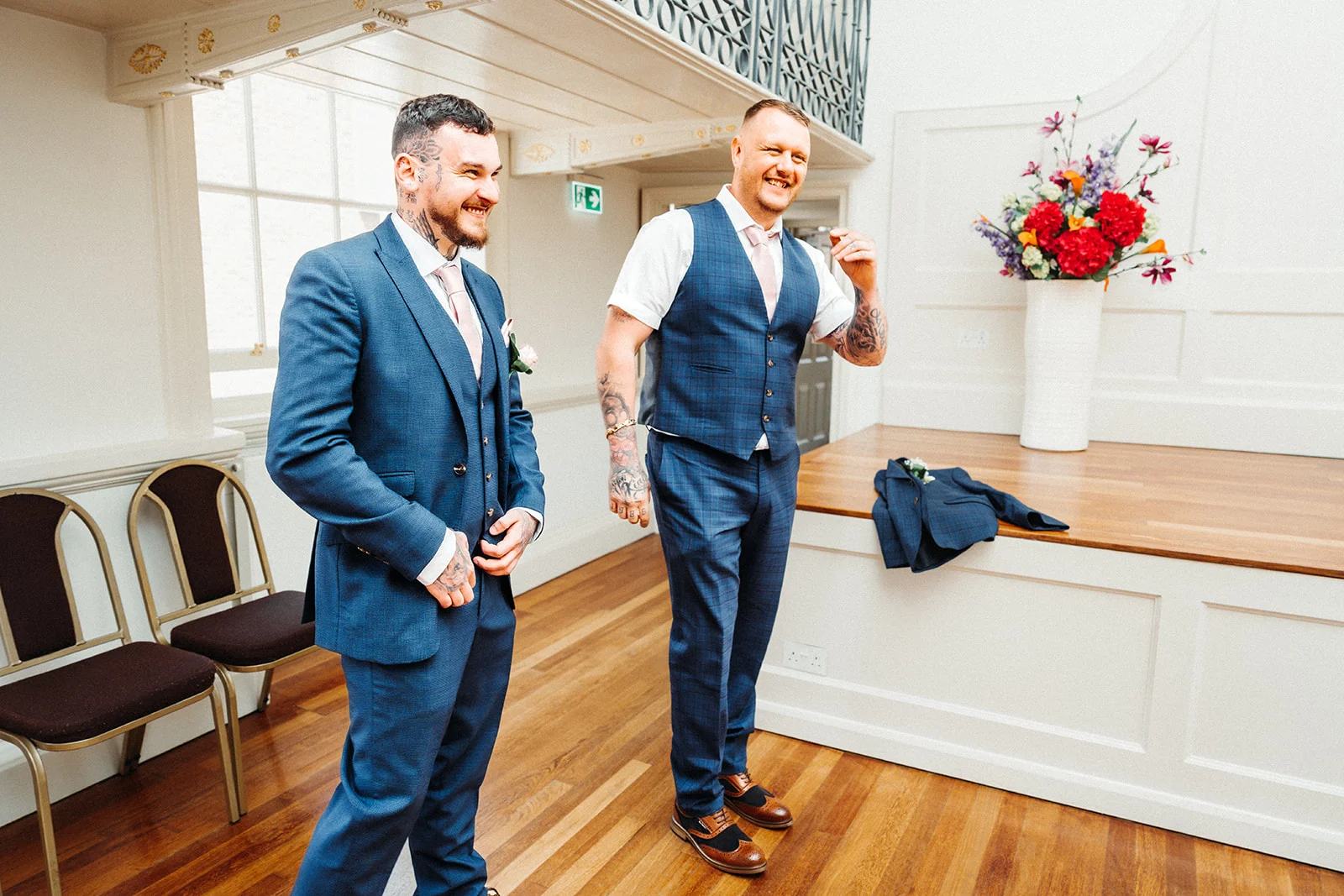Two grooms in matching blue suits share a joyful laugh while getting ready inside a bright, wood-floored venue adorned with floral arrangements.