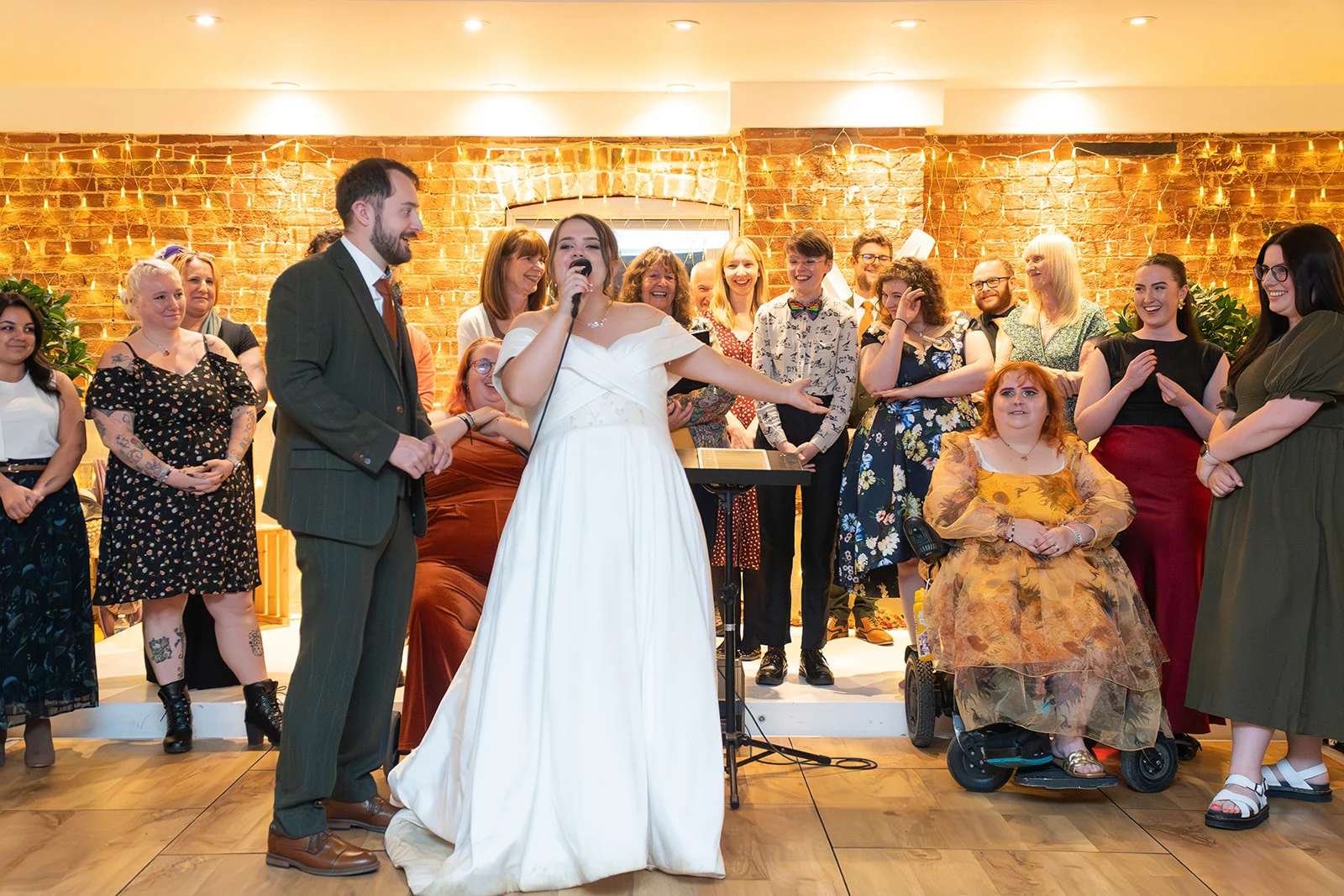 A bride in a white off-the-shoulder satin gown joyfully speaks into a microphone while standing next to her groom in a dark green suit, with smiling guests gathered in front of an exposed brick wall adorned with string lights.
