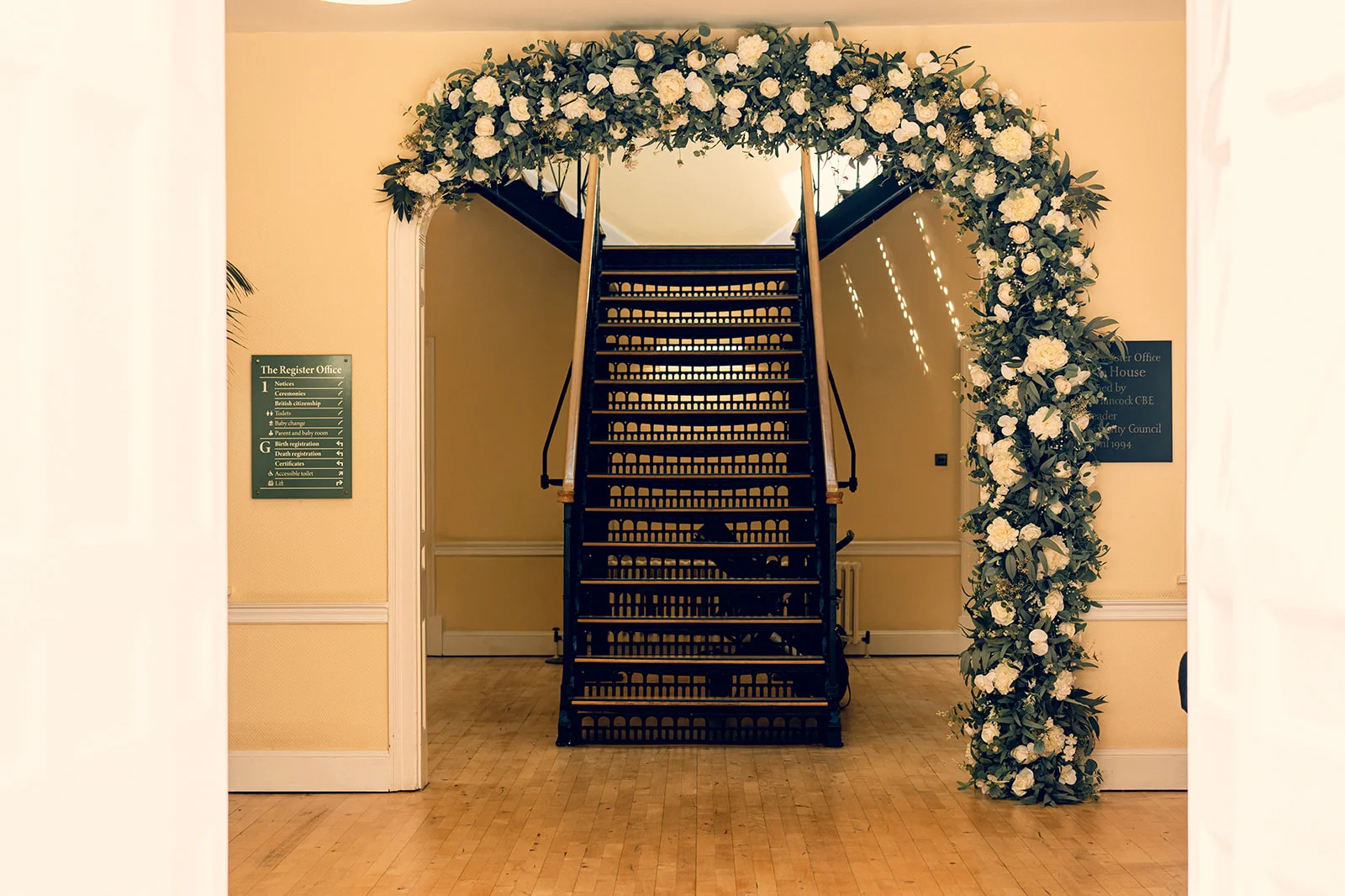 A grand central staircase inside a registry office is framed by a magnificent white and green floral arch featuring lush roses and eucalyptus foliage.