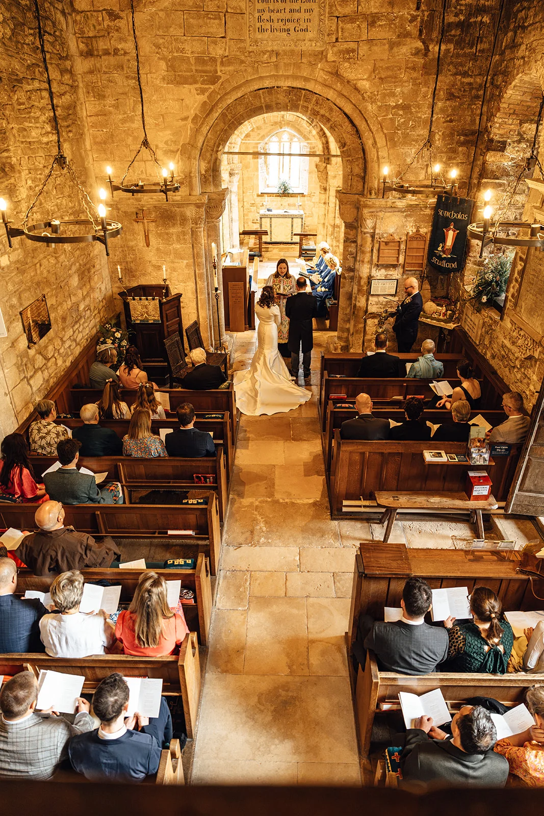 An elevated wide-angle view captures a wedding ceremony taking place inside a historic stone church with wooden pews and an arched chancel.