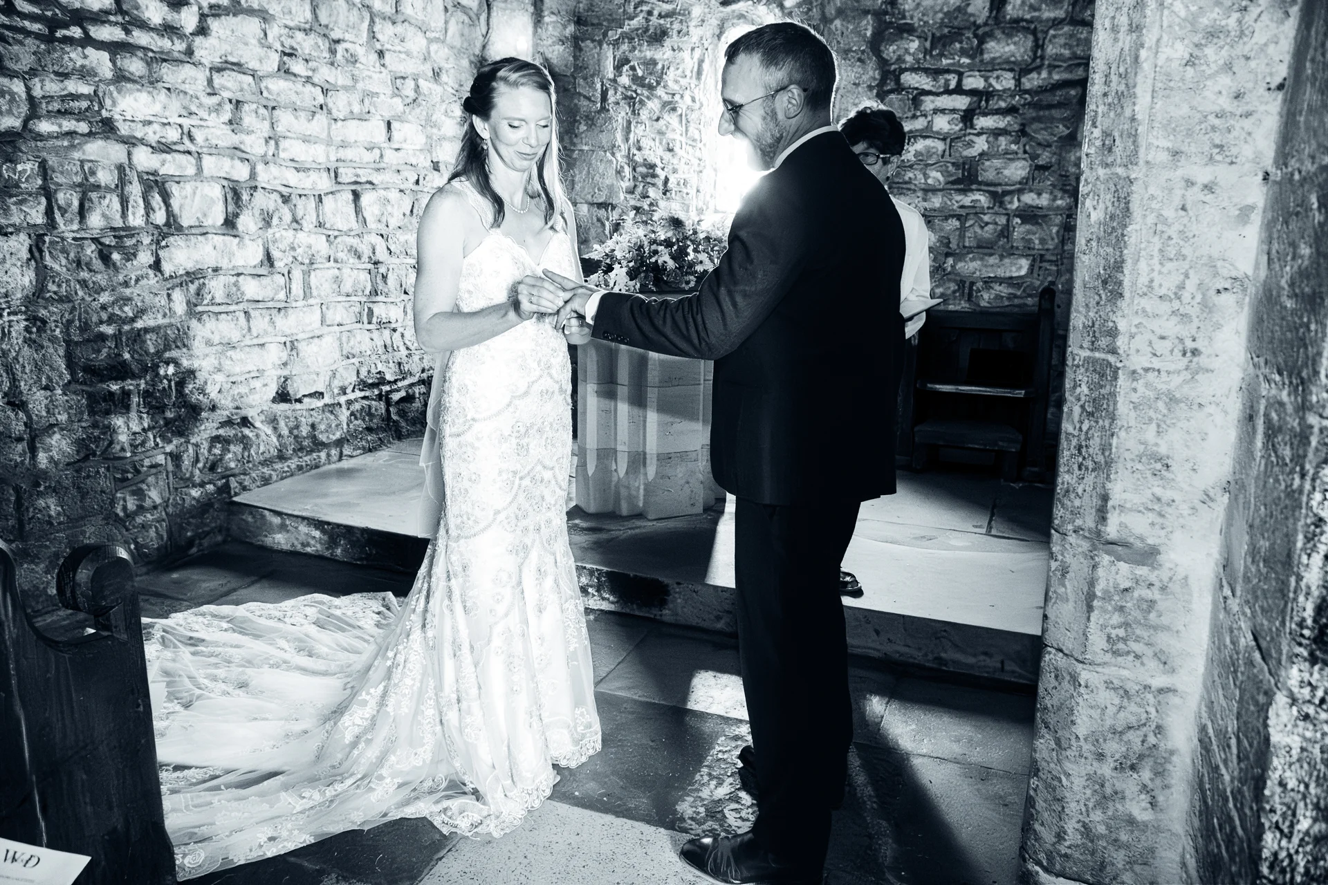 A groom places a ring on the bride's finger during their ceremony inside a historic stone church captured in dramatic black and white.