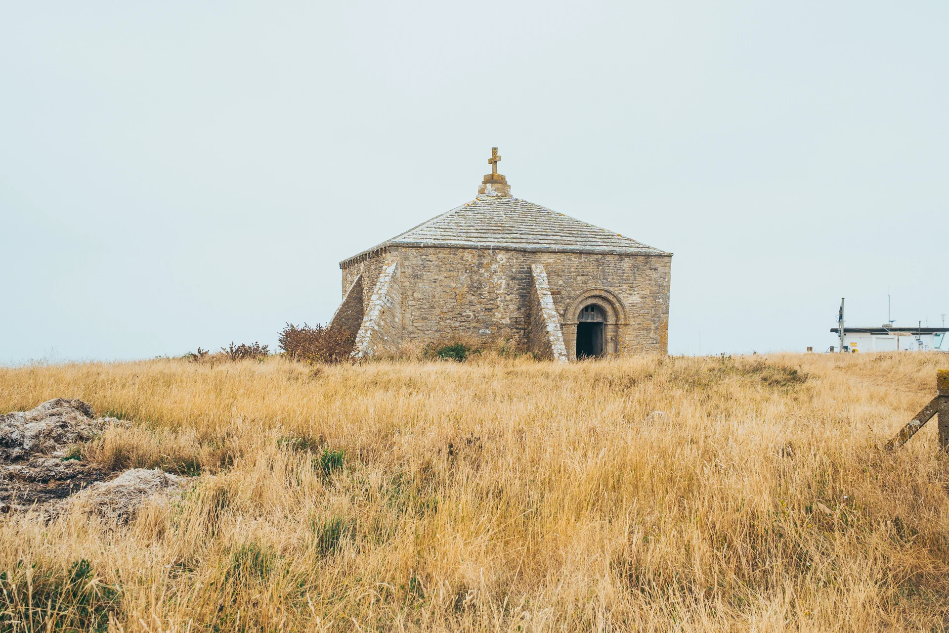 An ancient stone chapel with a cross on top sits amidst tall, golden grass under an overcast sky.