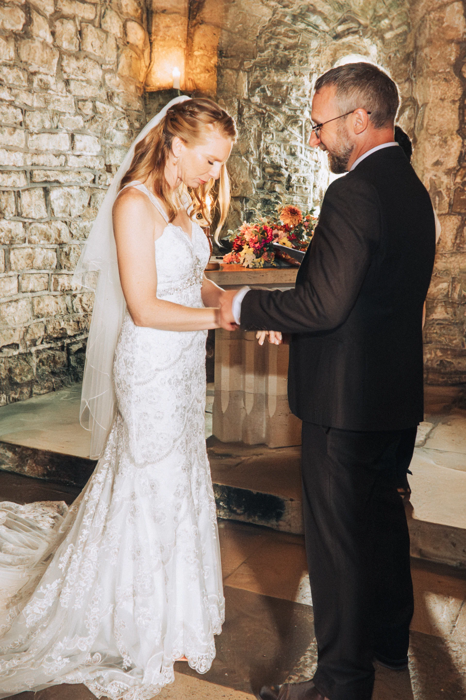 A bride and groom exchange rings during an intimate indoor ceremony inside a rustic stone venue with candlelight.