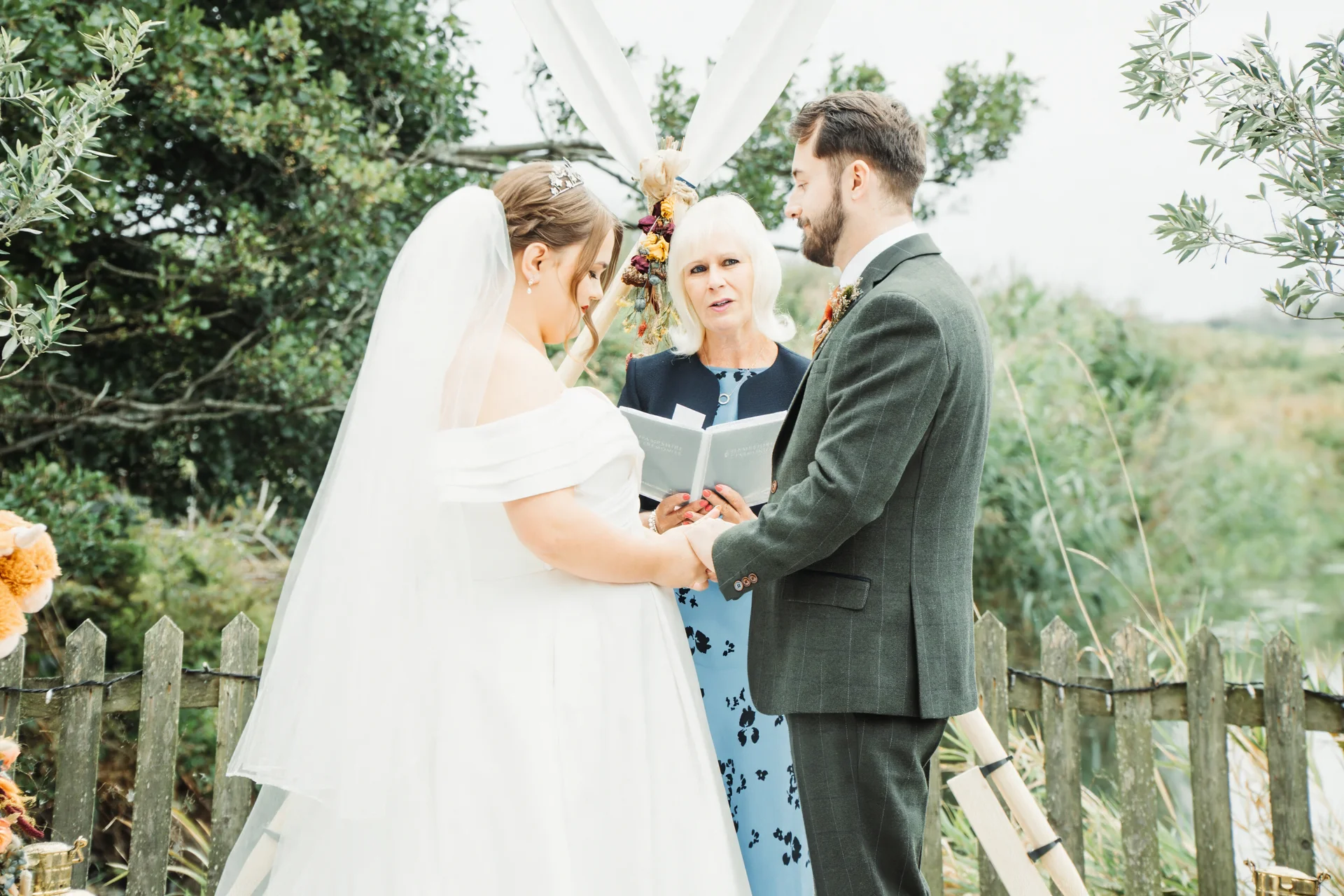 An outdoor wedding ceremony featuring a bride in an off-the-shoulder gown and veil holding hands with a groom in a green suit while a celebrant officiates.