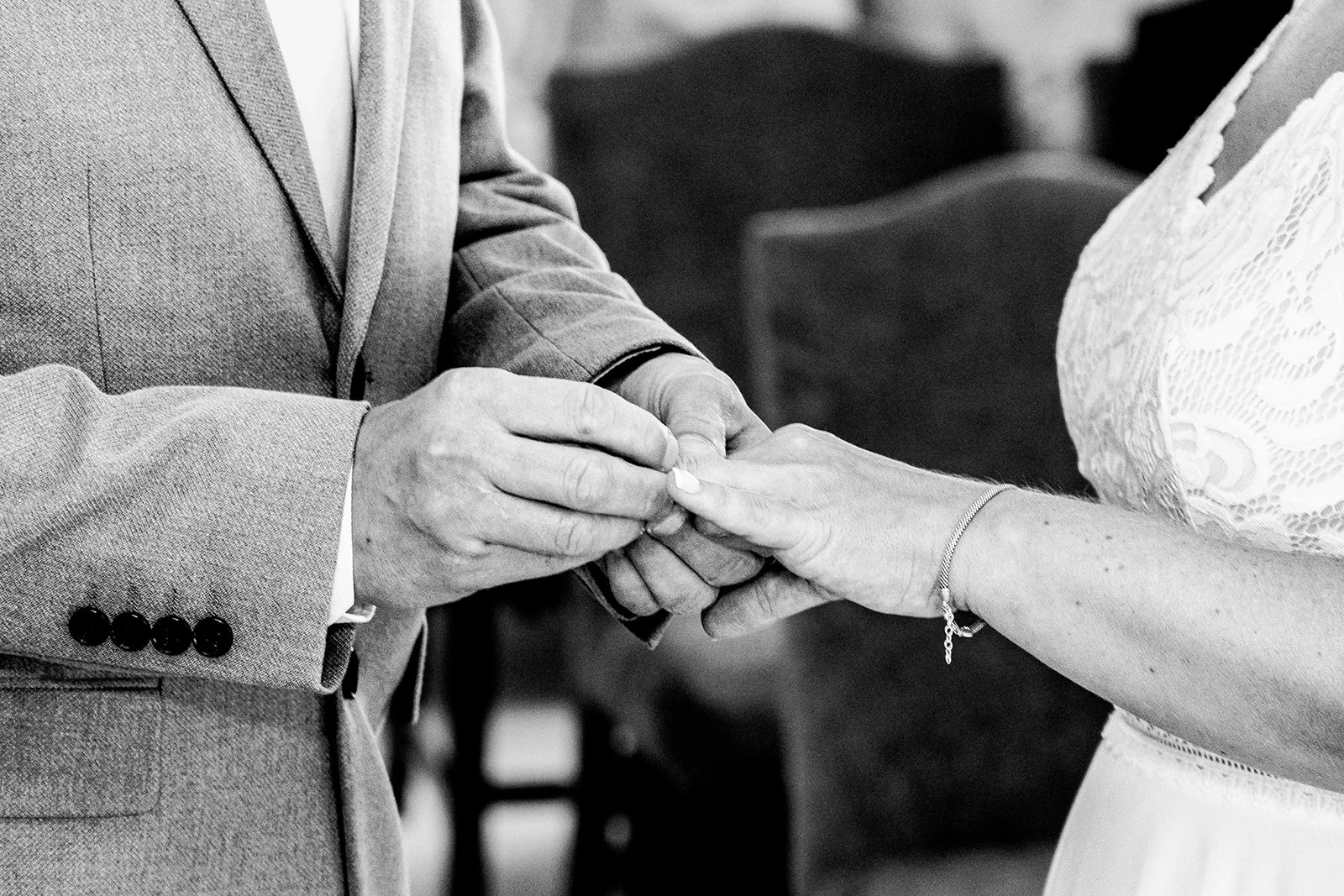 A close-up black and white photograph captures thegroom placing a wedding ring onto the bride's finger during the ceremony.