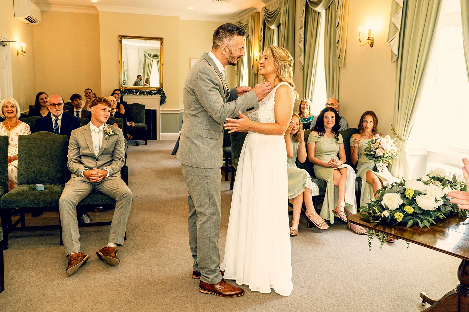 A groom in a grey suit and the bride in a white gown exchange joyful smiles during their indoor wedding ceremony.
