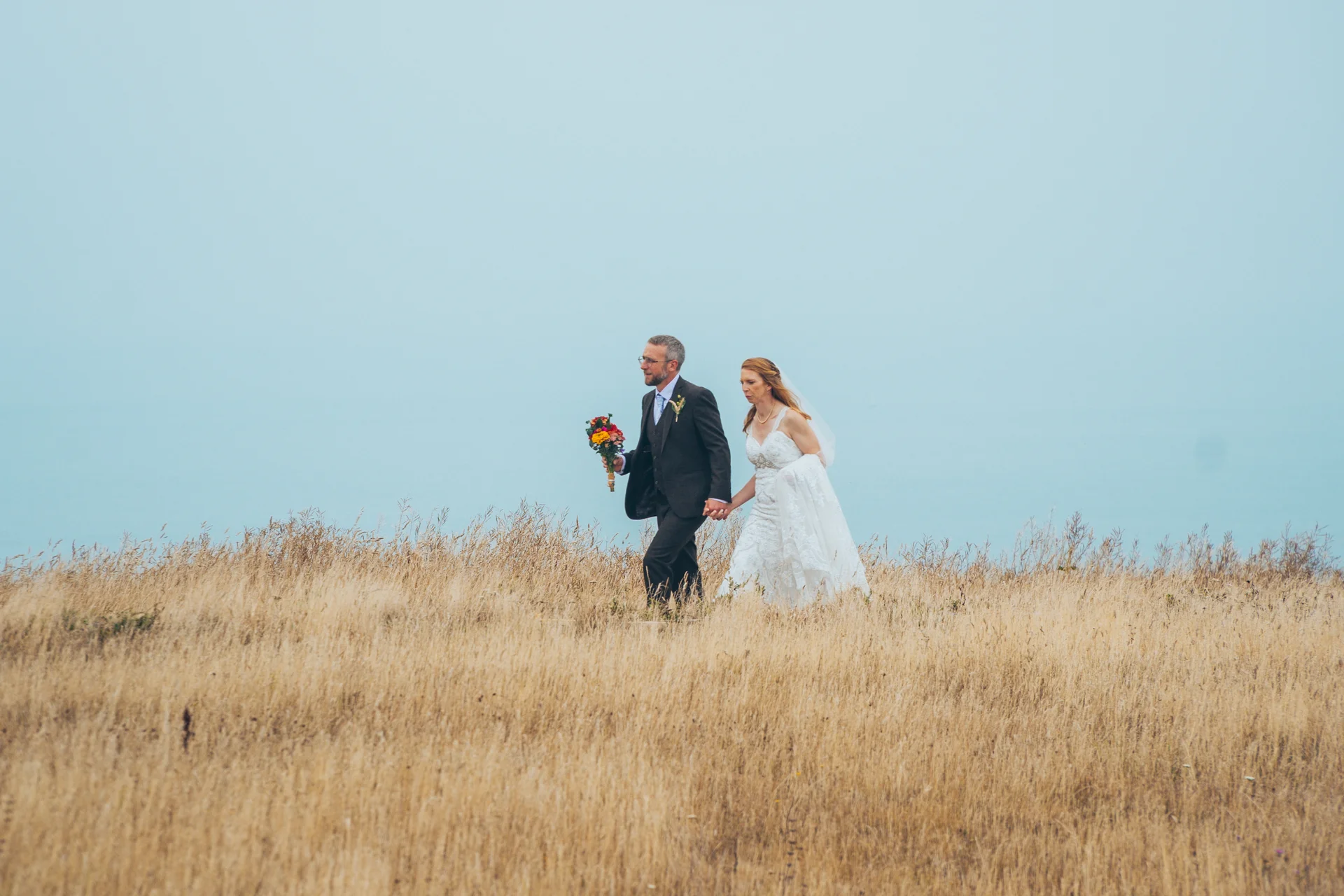 A newlywed couple holding hands and walking through a tall grass meadow beneath an expansive, muted sky.