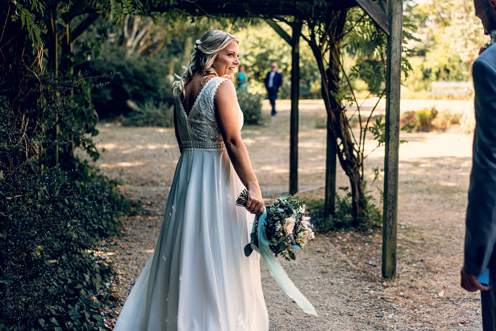 A smiling bride holding a bouquet with trailing ribbons looks back over her shoulder beneath a wooden pergola in a sunlit garden.