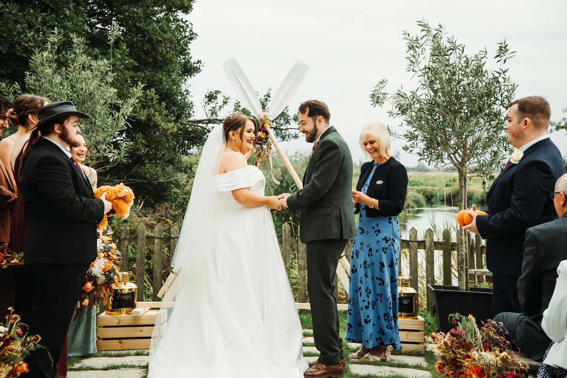 A joyful bride and groom exchange rings during an outdoor lakeside ceremony, surrounded by smiling wedding party members and guests.