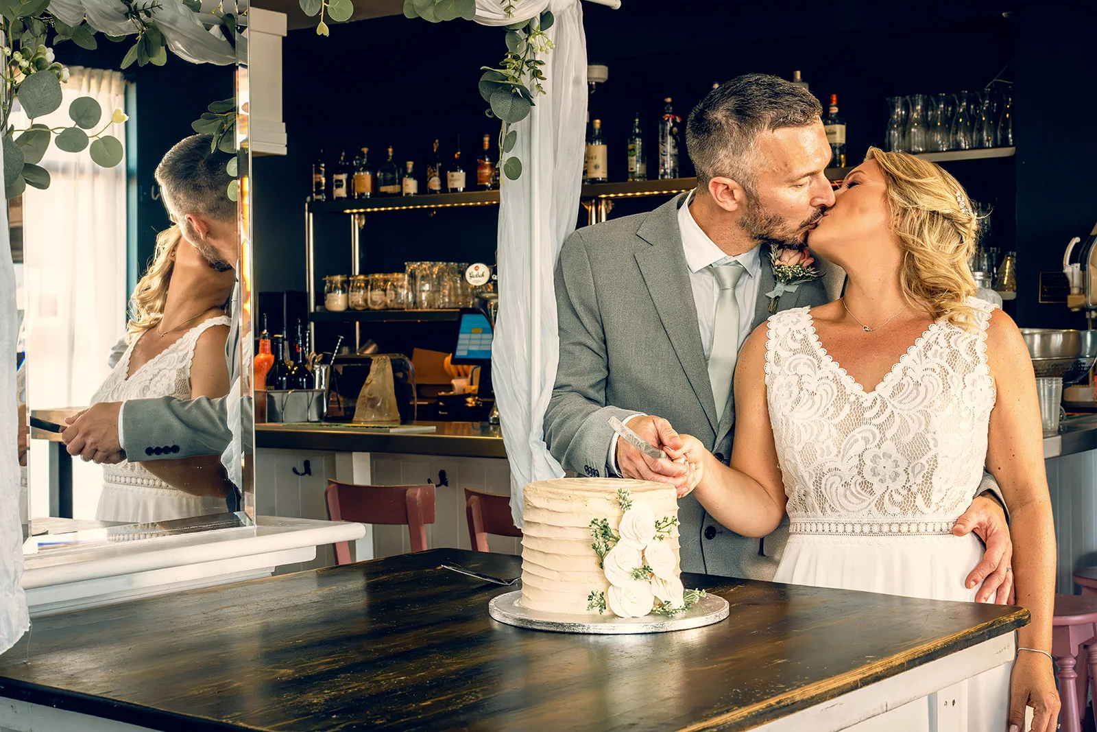 A bride and groom share a kiss while cutting their wedding cake together inside a pub venue, captured with a reflection in a nearby mirror.