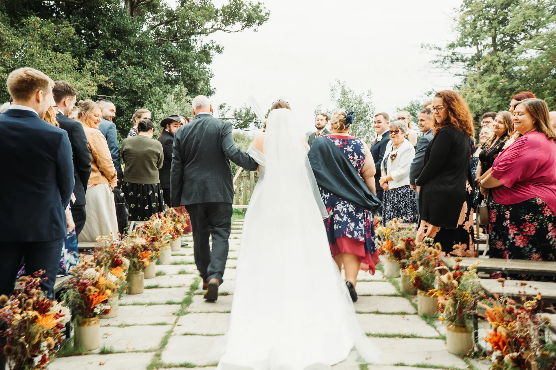 A bride walks down an outdoor stone-paved aisle lined with colourful autumn floral arrangements, escorted by her father as seated guests look on.