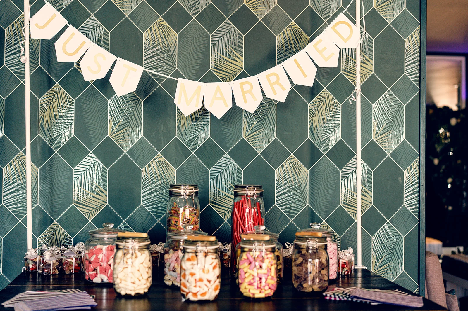 A vibrant pick and mix sweet table features glass jars filled with colorful confectionery underneath a just married banner against a patterned green wallpaper backdrop.